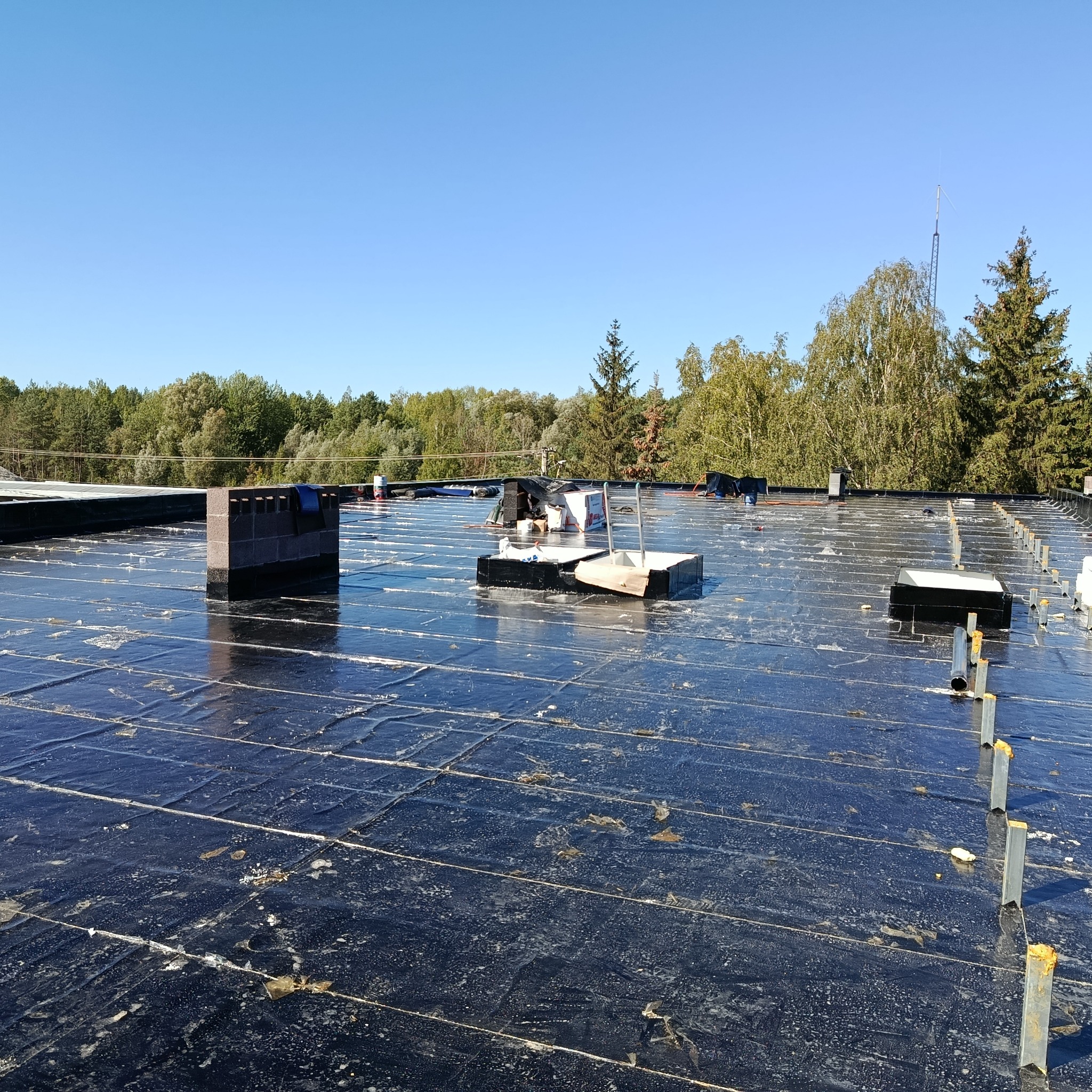 Czarna membrana dachowa z widocznymi elementami montażowymi i kominem w tle, w słoneczny dzień. Widok na zalesiony horyzont i wieżę telekomunikacyjną.