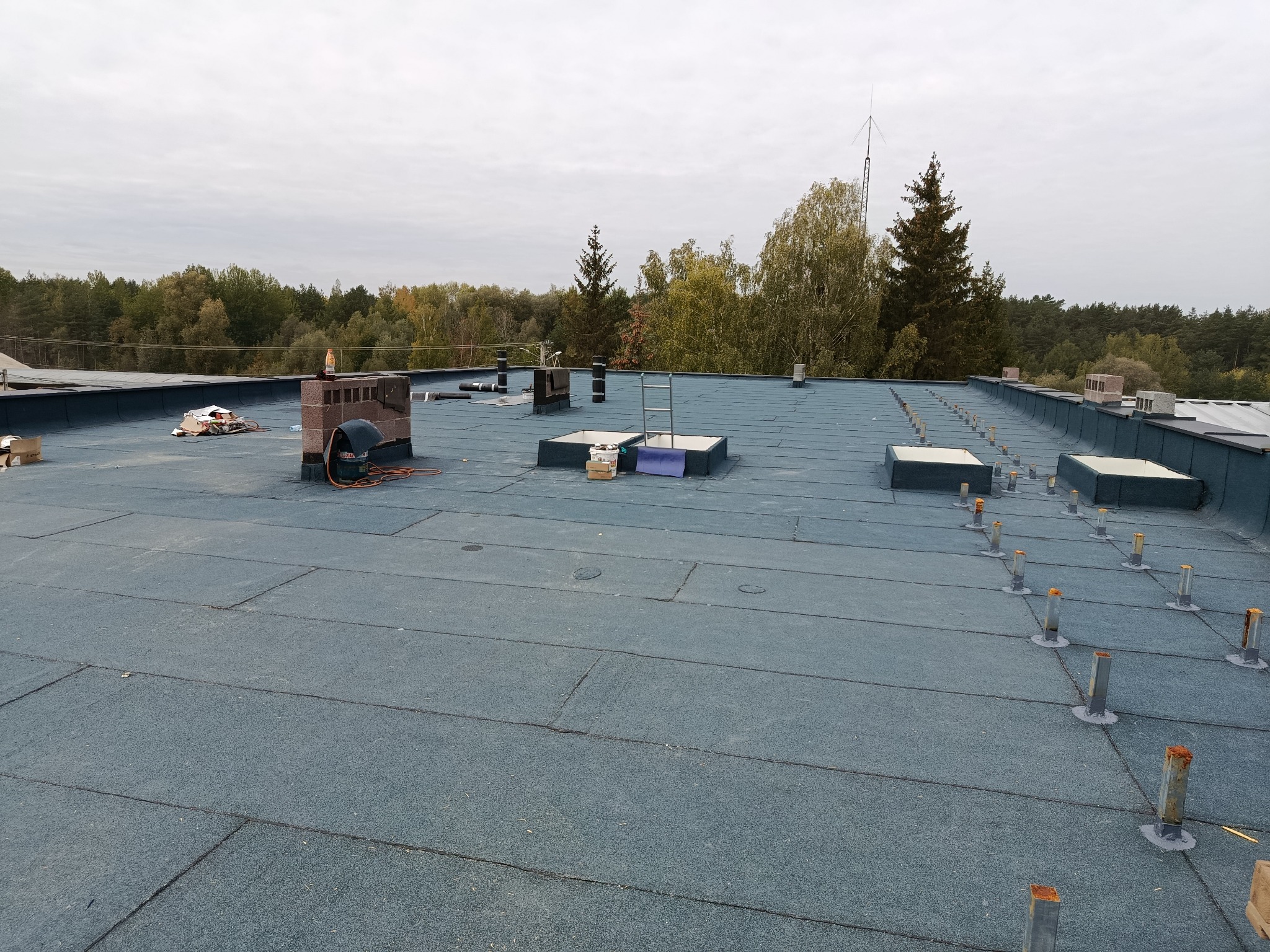 Płaski dach pokryty papą termozgrzewalną z kominem i elementami instalacji odgromowej na tle lasu i pochmurnego nieba. Widoczne narzędzia i materiały budowlane.