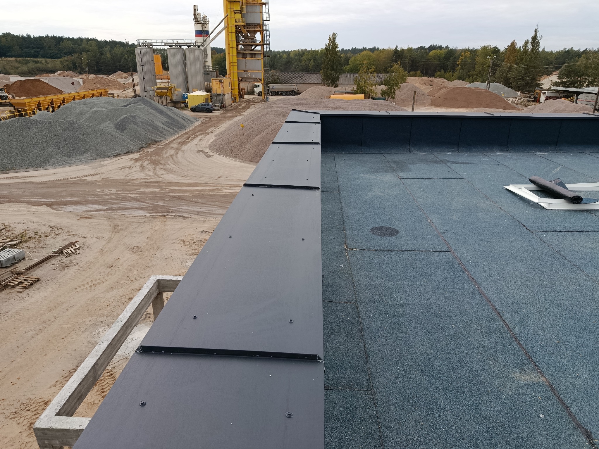 Szczegółowe ujęcie płaskiego dachu pokrytego papą termozgrzewalną, z widocznymi obróbkami blacharskimi i elementami mocującymi. W tle hałda piasku i żwiru.