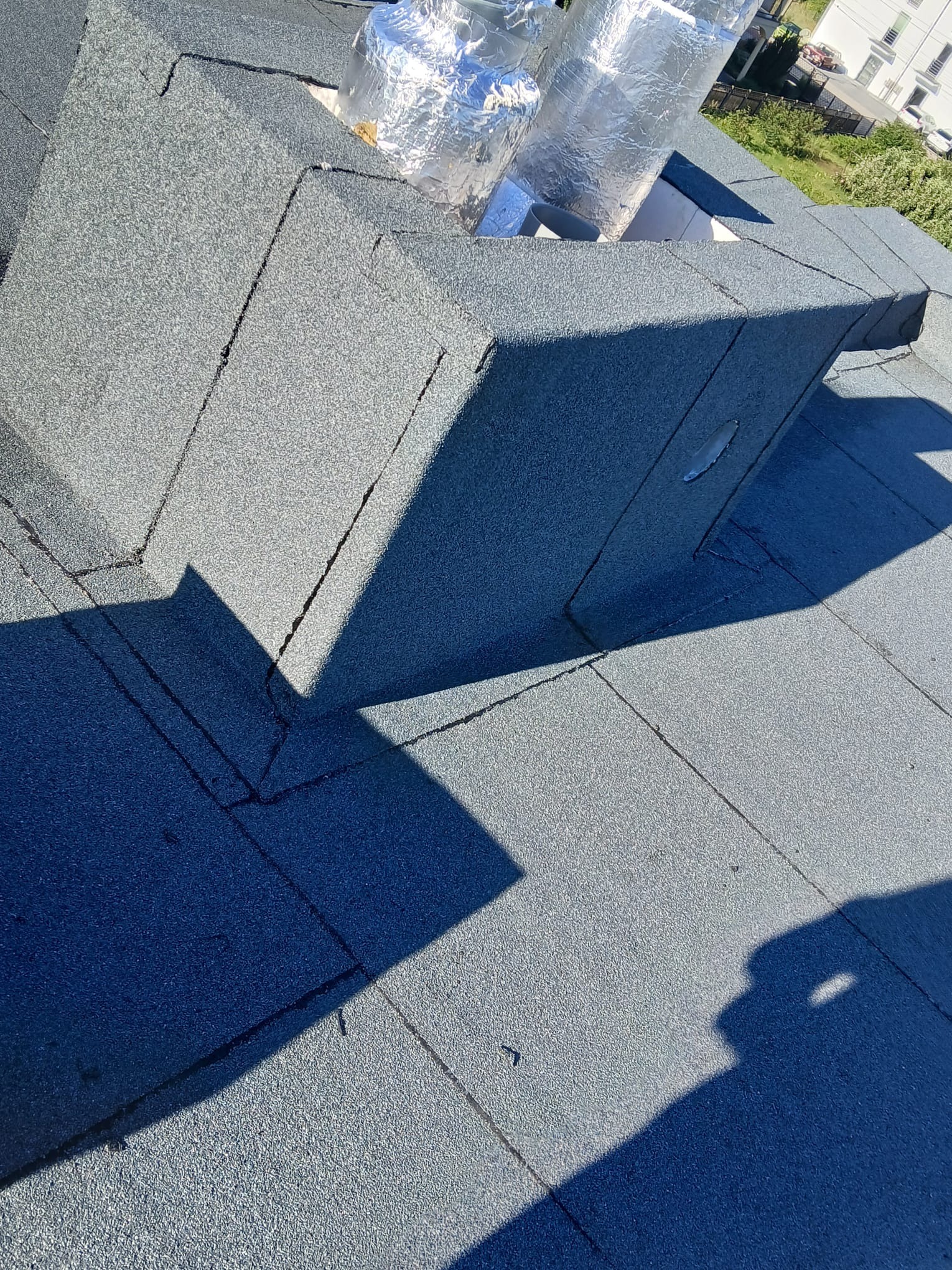 Szczegółowe ujęcie płaskiego dachu pokrytego papą z widocznym kominem wentylacyjnym zabezpieczonym folią aluminiową. Ciemne cienie podkreślają strukturę pokrycia.