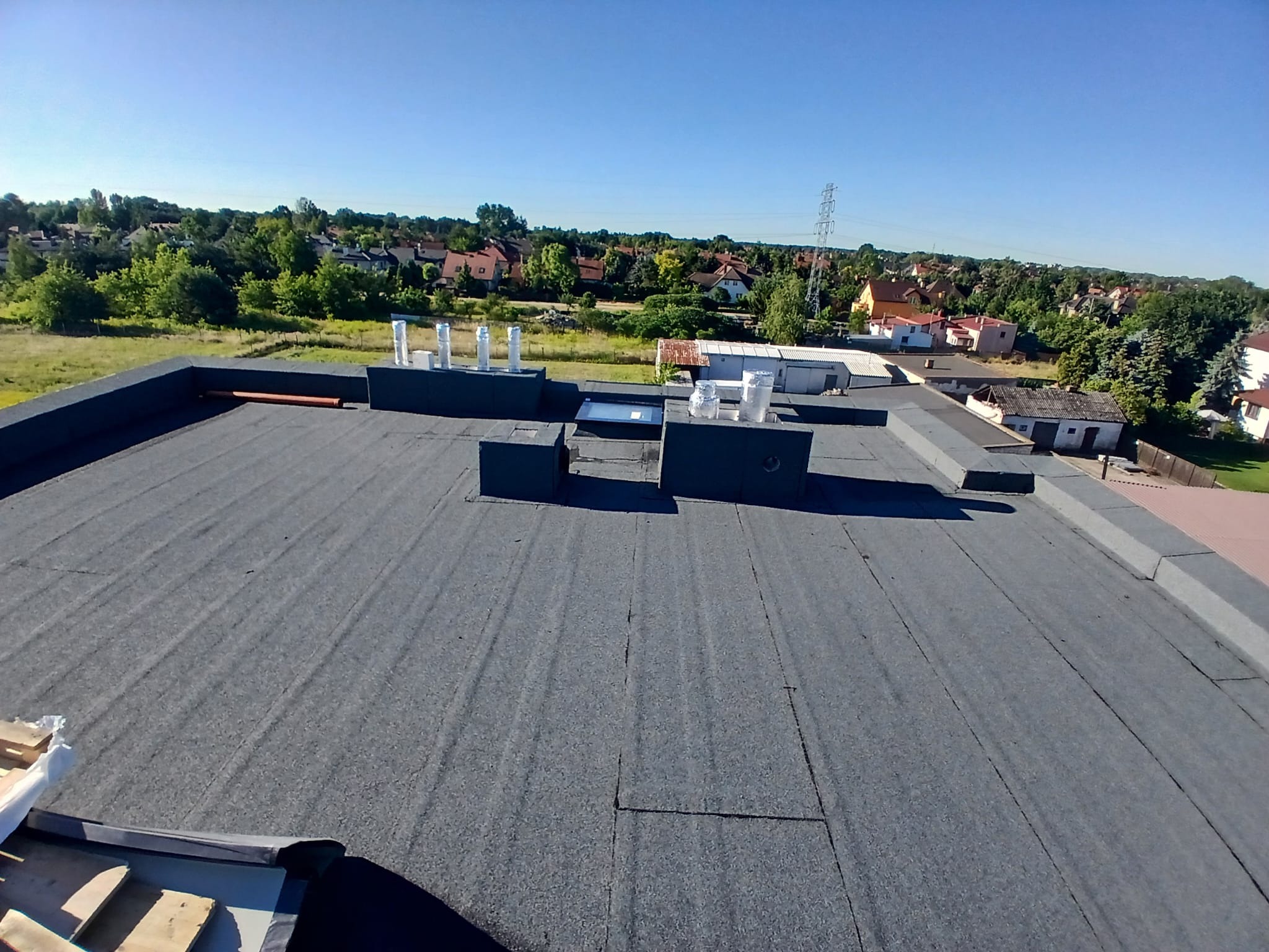 Szary, płaski dach pokryty papą termozgrzewalną z kominami wentylacyjnymi i świetlikiem. W tle zabudowa jednorodzinna i zieleń w słoneczny dzień.