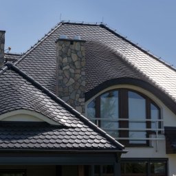 PIOTR SZCZUTKOWSKI - Elewacja domu z dachem pokrytym czarną dachówką łuskową i kamiennym kominem. Widoczne okno balkonowe z balustradą. Dach po renowacji.
