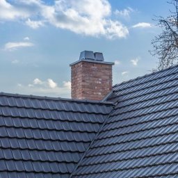 PIOTR SZCZUTKOWSKI - Szary dach z ceramicznej dachówki, ceglany komin z metalowym wykończeniem na tle błękitnego nieba z chmurami i gałęzi drzewa.