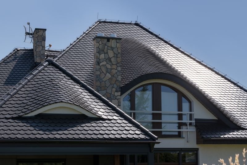 Elewacja domu z dachem pokrytym czarną dachówką łuskową i kamiennym kominem. Widoczne okno balkonowe z balustradą. Dach po renowacji.