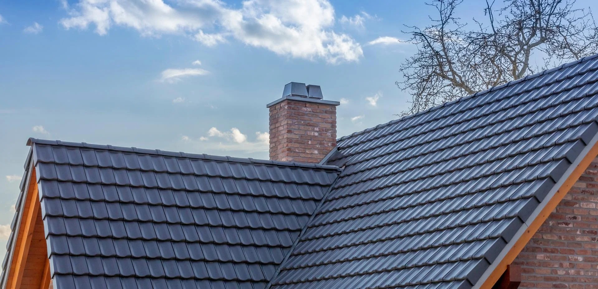 Szary dach z ceramicznej dachówki, ceglany komin z metalowym wykończeniem na tle błękitnego nieba z chmurami i gałęzi drzewa.