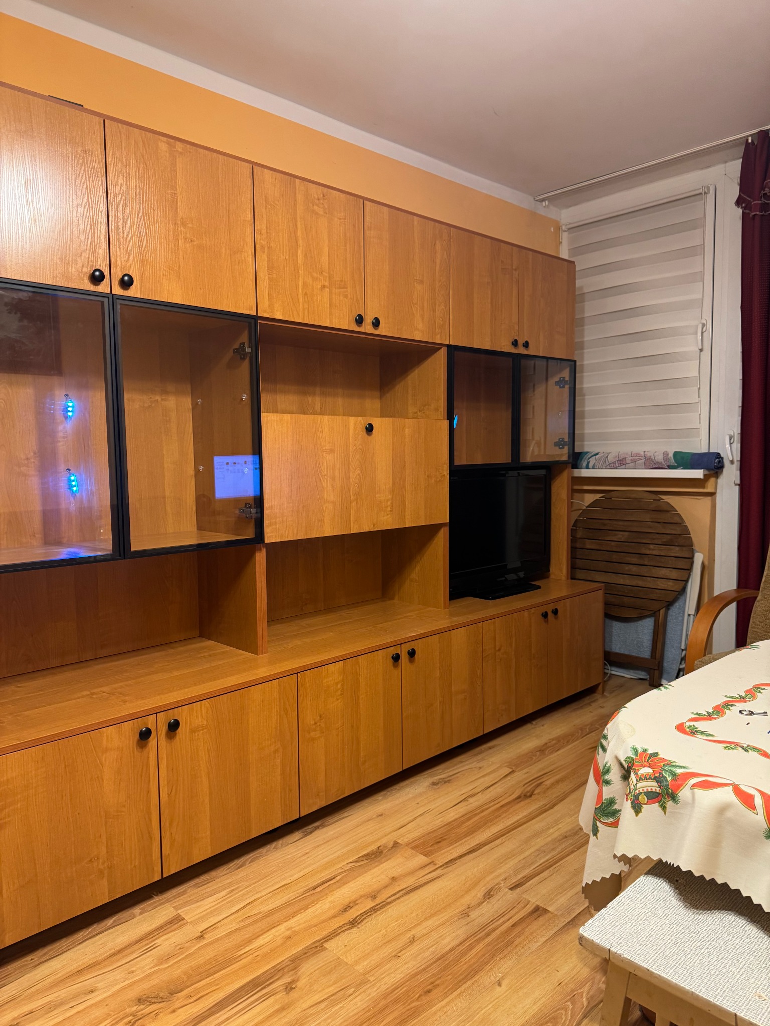 Drewniana meblościanka z czarnymi akcentami i przeszklonymi witrynami, w tle okno z roletami i stół z świątecznym obrusem. Ciepłe oświetlenie wnętrza.