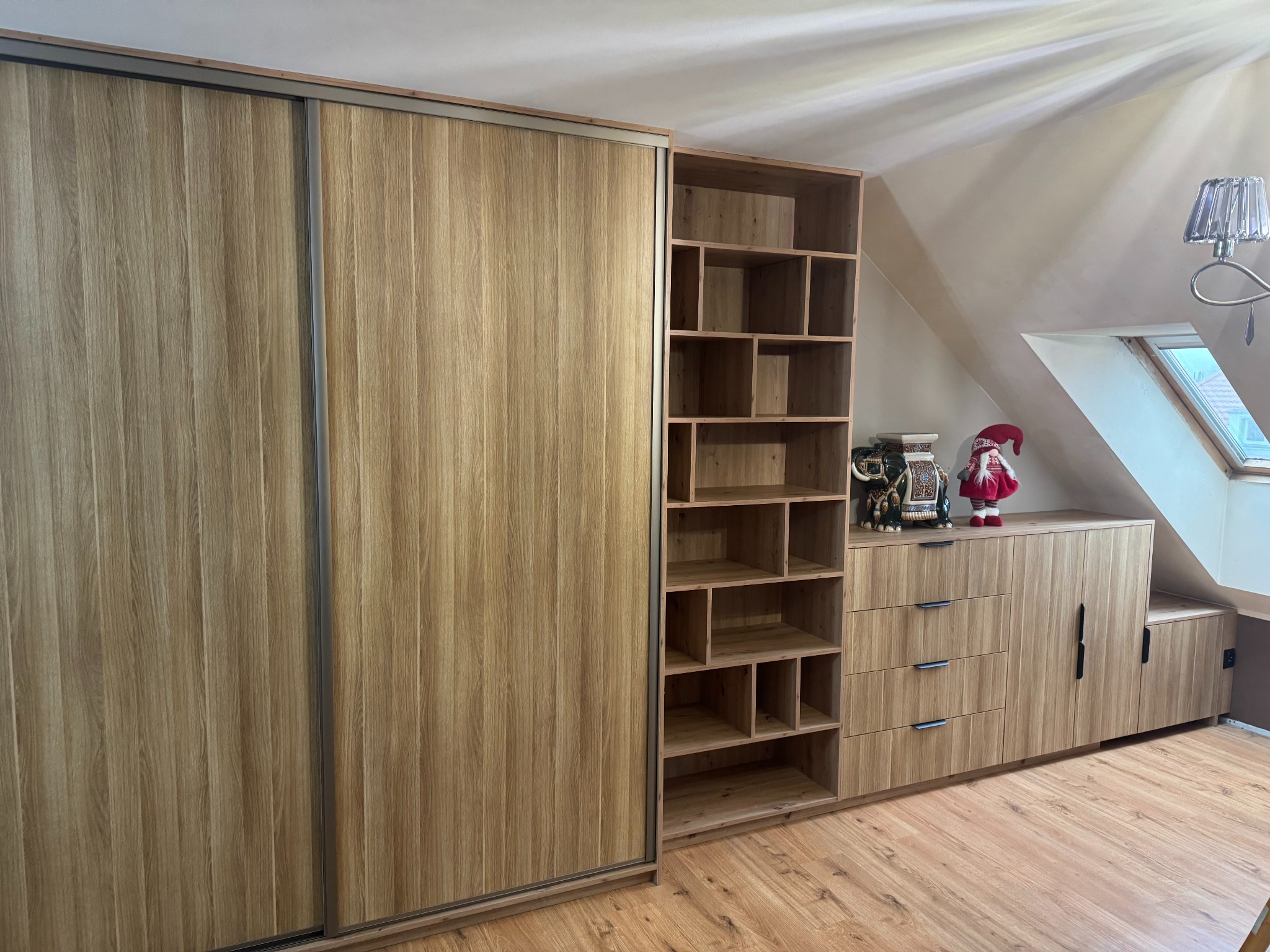 Drewniana szafa przesuwna z półkami w zabudowie pod skosem. Jasne drewno, minimalistyczne uchwyty, oświetlenie sufitowe. Aranżacja wnętrza.