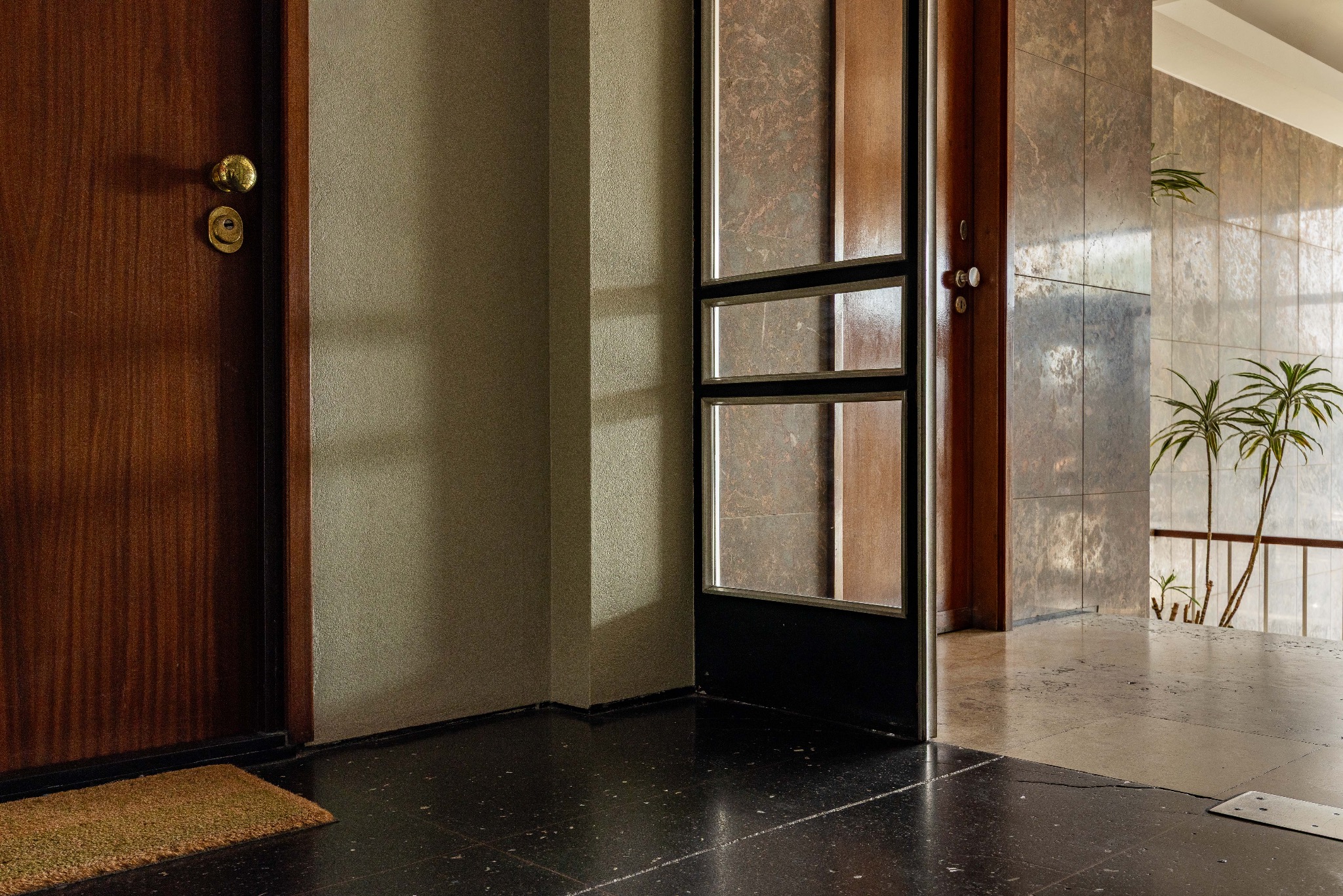 Kadr z wnętrza: ciemne drzwi z mosiężną gałką, fragment ściany, przeszklone drzwi z czarną ramą, marmurowe płytki i rośliny w tle. Ujęcie z niskiego kąta.