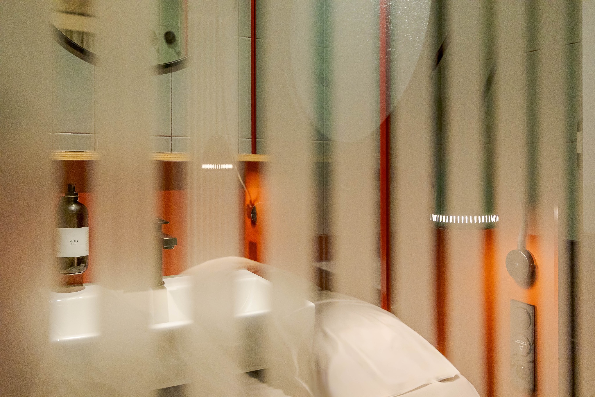 Nowoczesna łazienka z umywalką, dozownikiem mydła i pionowymi elementami dekoracyjnymi w odcieniach czerwieni i zieleni, widziana przez lekko zamgloną szybę.