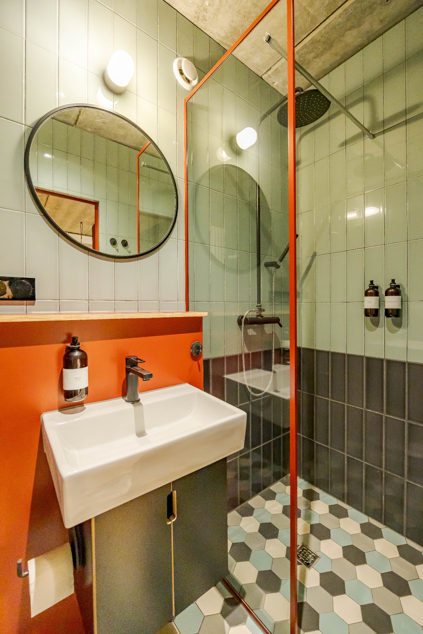 Nowoczesna łazienka z kabiną prysznicową oddzieloną szkłem w pomarańczowej ramie, umywalką i okrągłym lustrem na tle pistacjowych płytek.