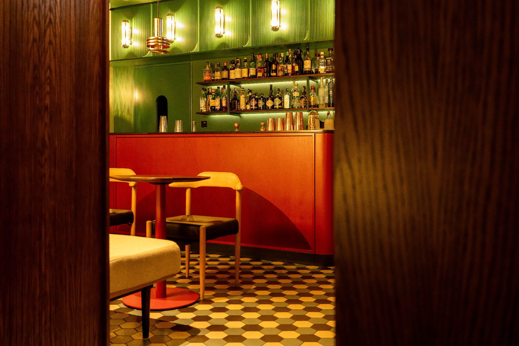 Stylowy bar z zielonymi ścianami, czerwonym blatem i półkami z butelkami. W tle widoczne stoliki z krzesłami i wzorzysta podłoga. Ujęcie zza drewnianej framugi.