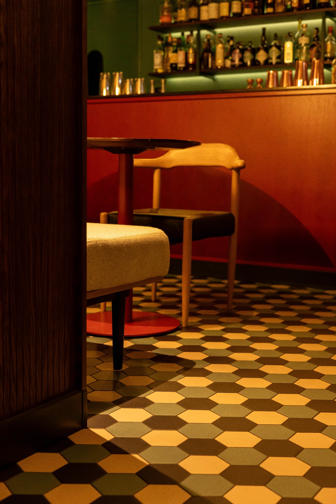 Stylowe wnętrze baru z heksagonalną podłogą, krzesłem i stołem na czerwonej podstawie. W tle półki z butelkami i barmańskimi akcesoriami, oświetlone zielonym światłem.
