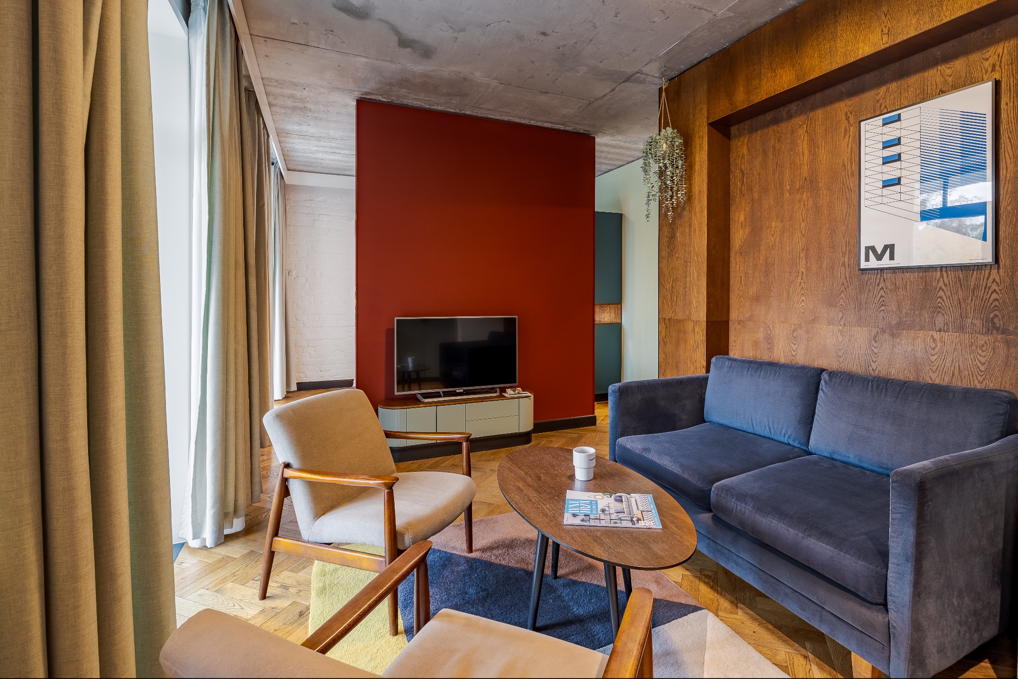 Stylowe wnętrze salonu z drewnianą boazerią, szarą sofą i fotelami w stylu retro. Na ścianie grafika w ramie i wisząca roślina. Widoczny telewizor i stolik kawowy.