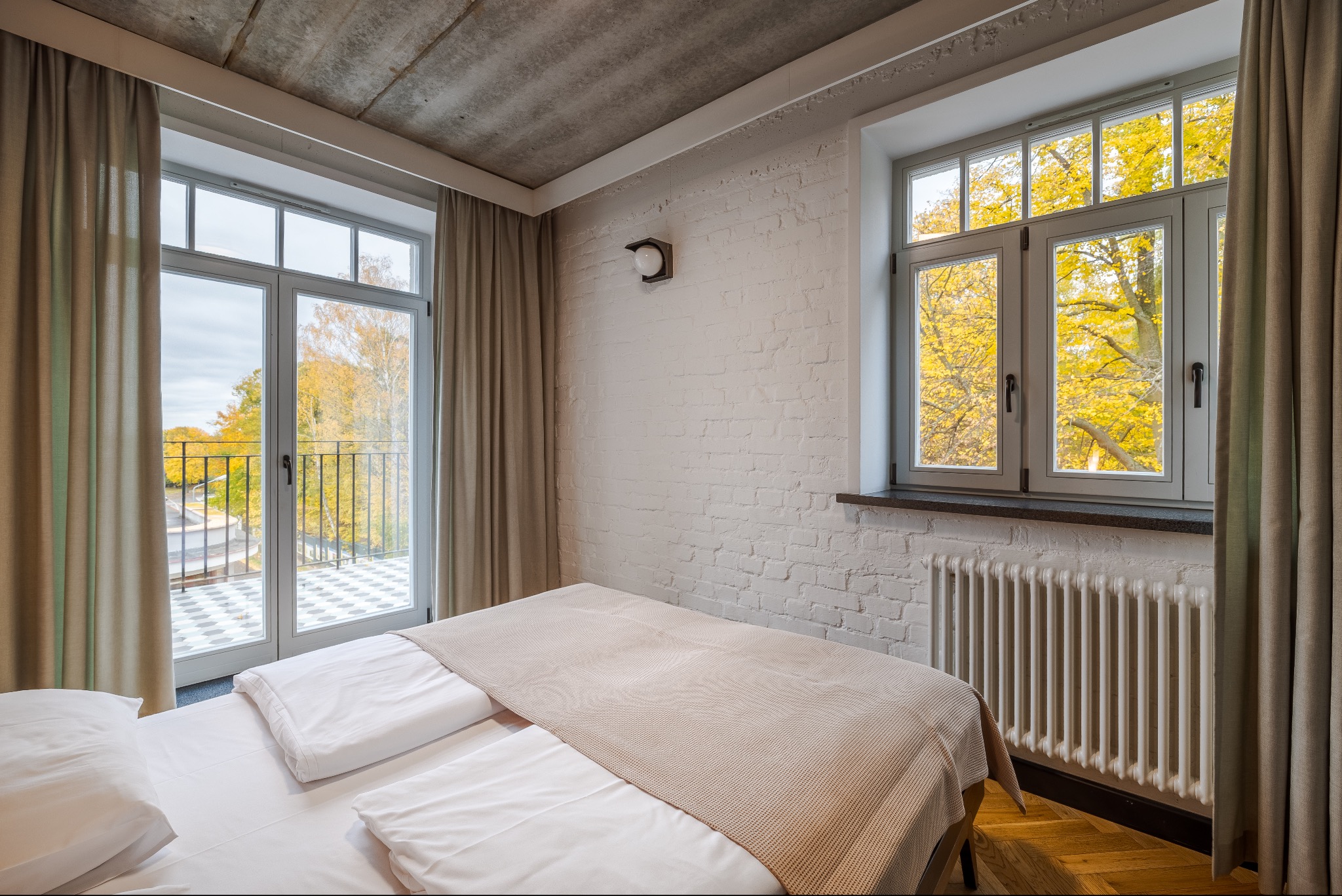 Jasna sypialnia z widokiem na jesienny krajobraz przez okno balkonowe i mniejsze okno. Biała cegła, drewniana podłoga, beżowe zasłony i betonowy sufit.