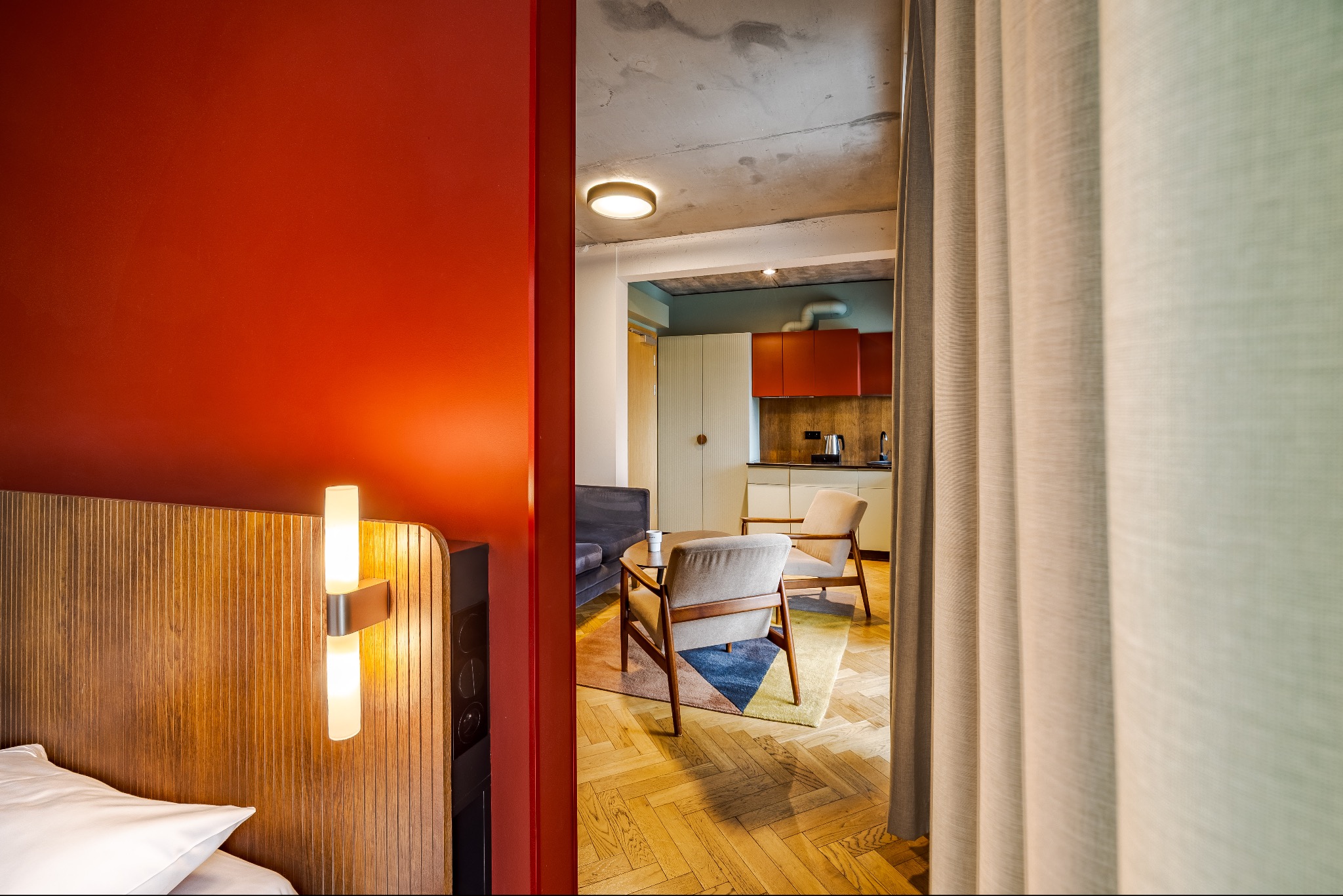 Nowoczesne wnętrze apartamentu z widokiem na salon i kuchnię, widziane zza zasłony. Czerwona ściana i drewniane panele z lampą przy łóżku w kadrze.