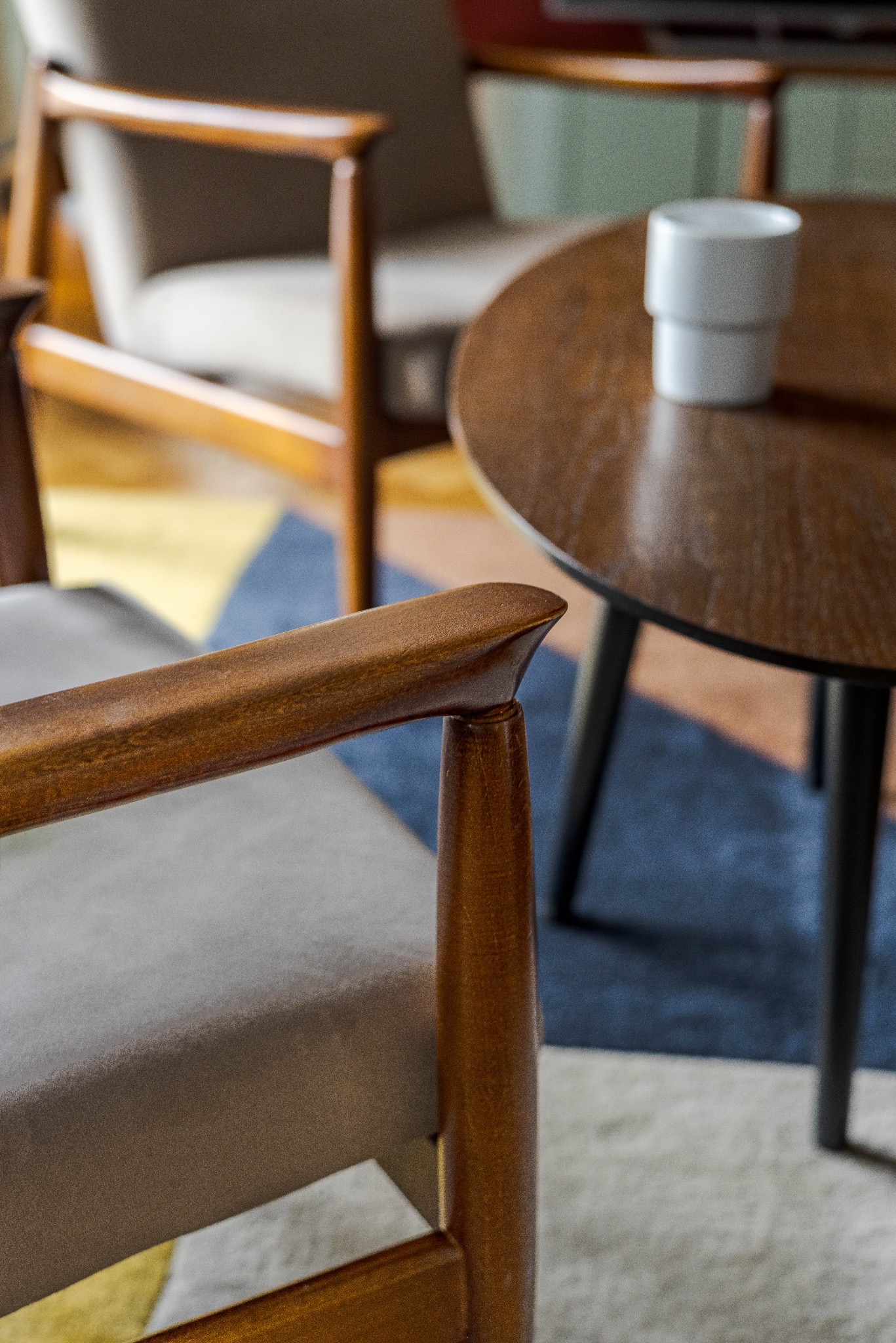 Detal drewnianego fotela w stylu retro z okrągłym stolikiem i kubkiem w tle. Ujęcie z bliska, podkreślające fakturę drewna i tkaniny obiciowej.