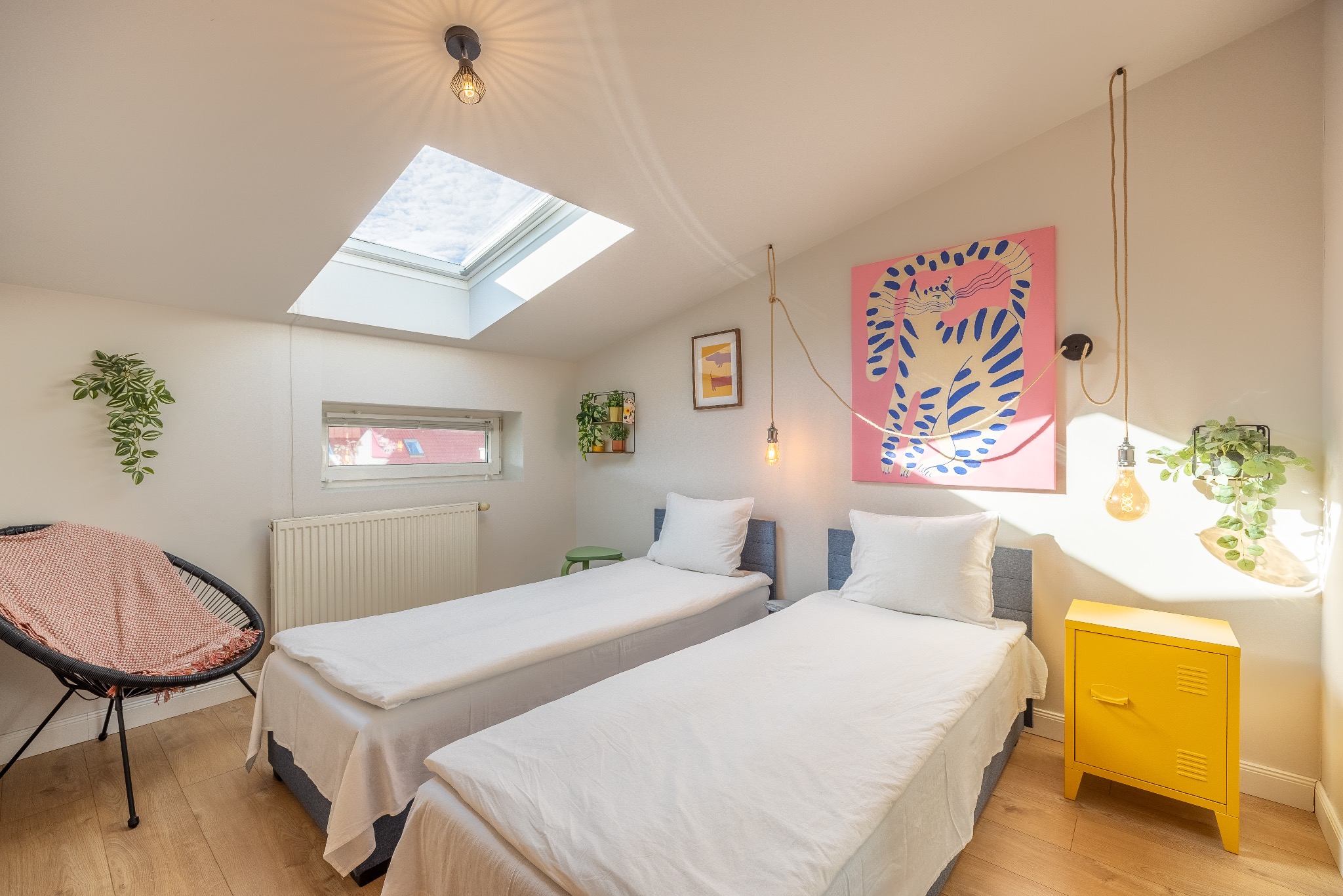 Jasna sypialnia na poddaszu z dwoma łóżkami, oknem dachowym i dekoracjami w stylu boho: lampy na sznurze, grafika z kotem i rośliny doniczkowe. Przytulne wnętrze.
