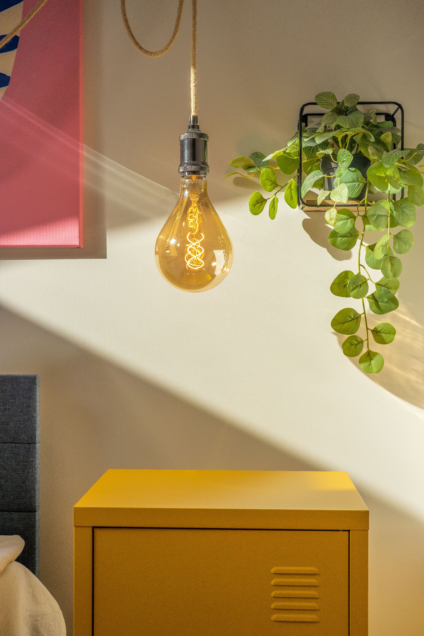 Żółta szafka nocna z lampą w stylu retro i rośliną w metalowej ramce na ścianie. Ujęcie w ciepłych barwach, z wyraźnym światłocieniem.