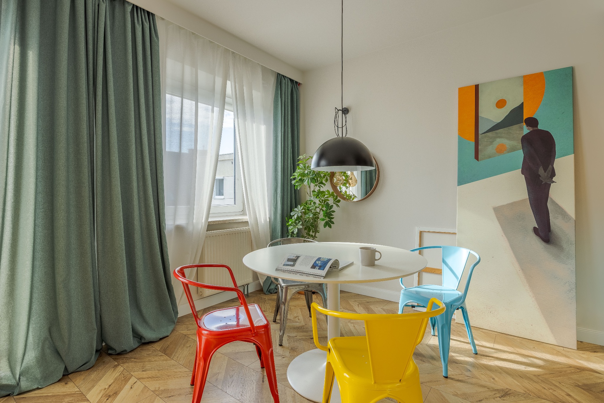 Jasne wnętrze jadalni z okrągłym stołem, kolorowymi krzesłami i nowoczesnym obrazem. Parkiet w jodełkę, zielone zasłony i roślina doniczkowa dodają przytulności.