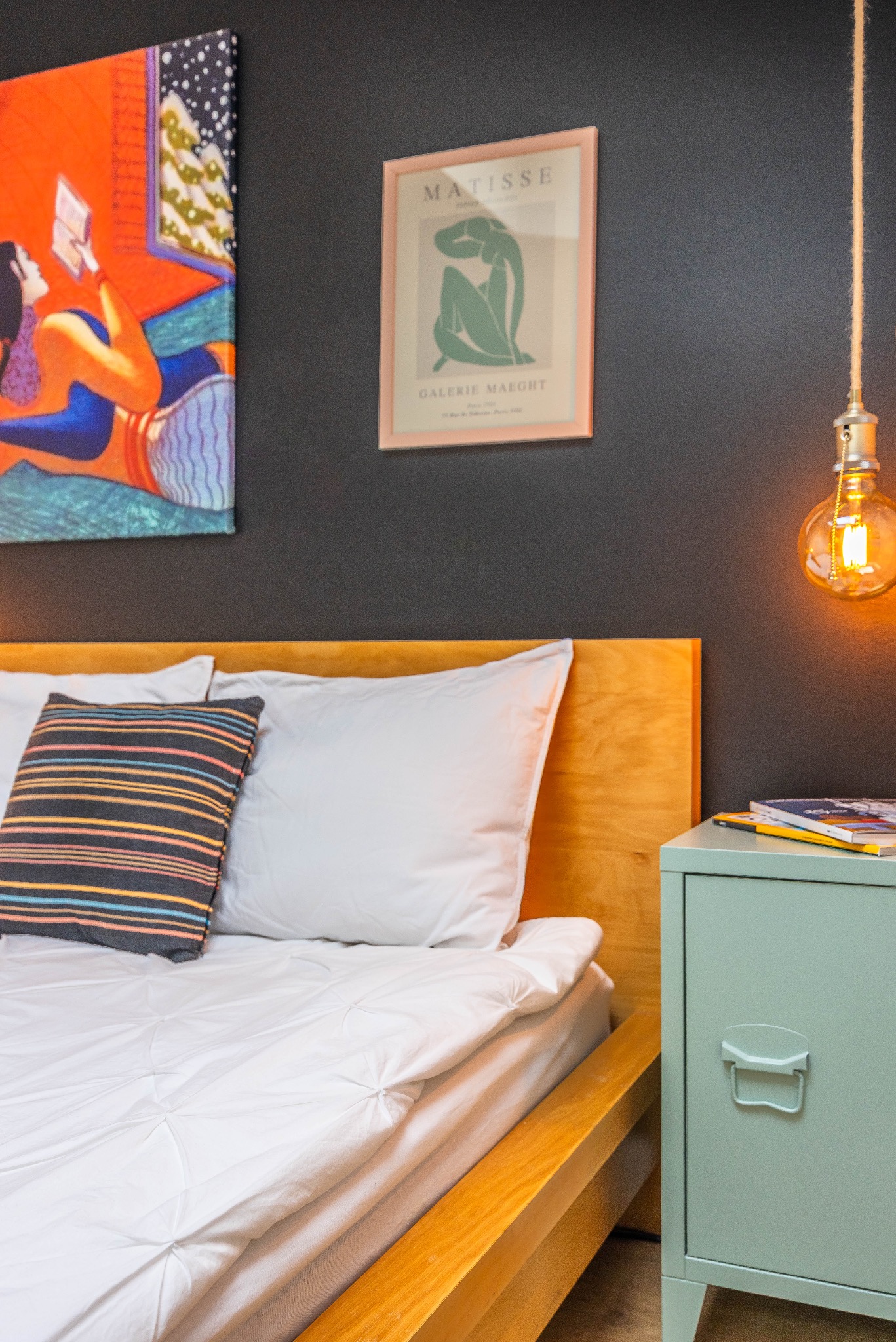 Sypialnia z drewnianym łóżkiem, białą pościelą i szafką nocną w odcieniu mięty. Na ścianie wiszą obrazy i lampa z żarówką Edisona.