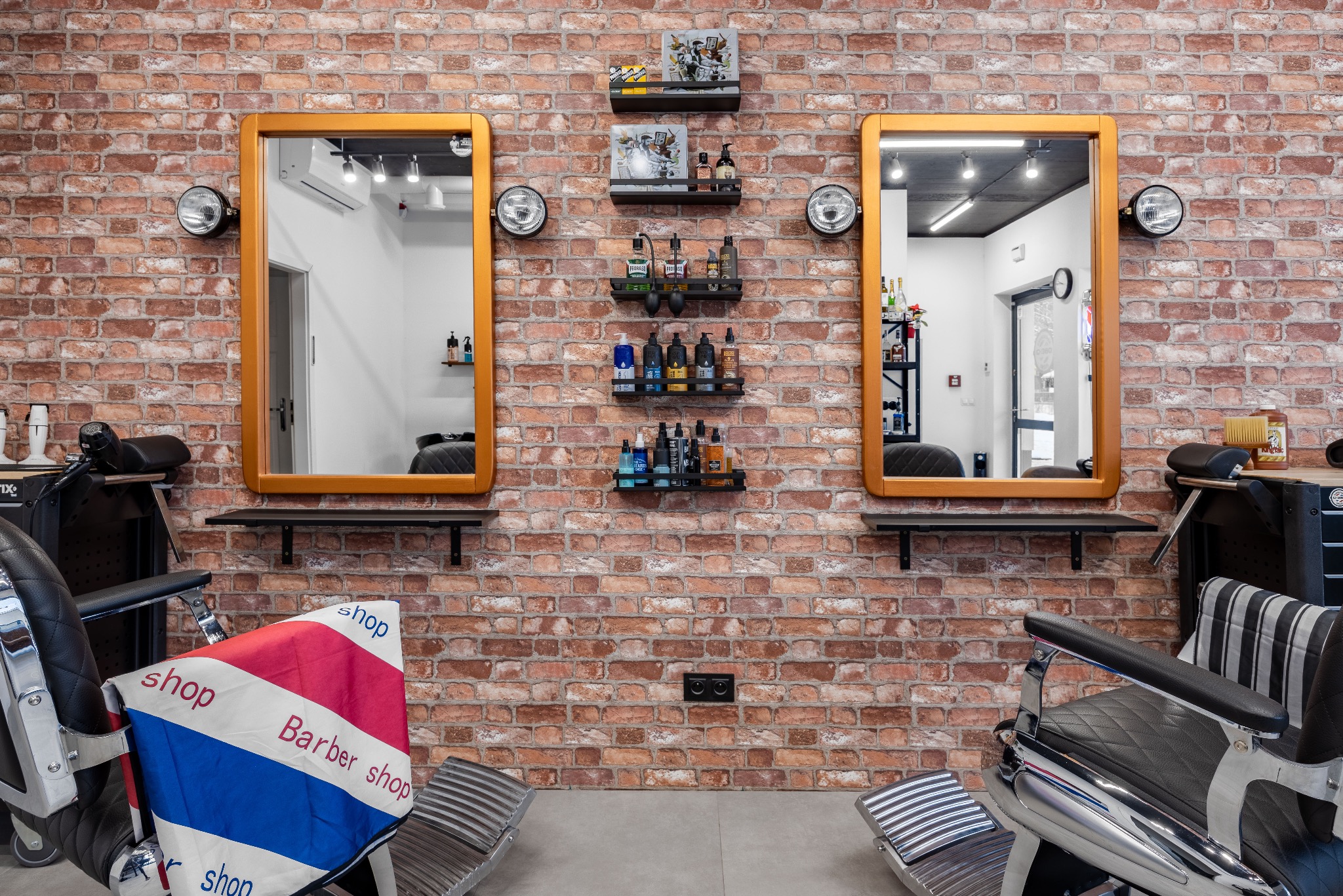 Wnętrze barber shopu z ceglaną ścianą, dwoma lustrami z oświetleniem i półkami z kosmetykami. Widoczne fotele fryzjerskie i akcesoria.