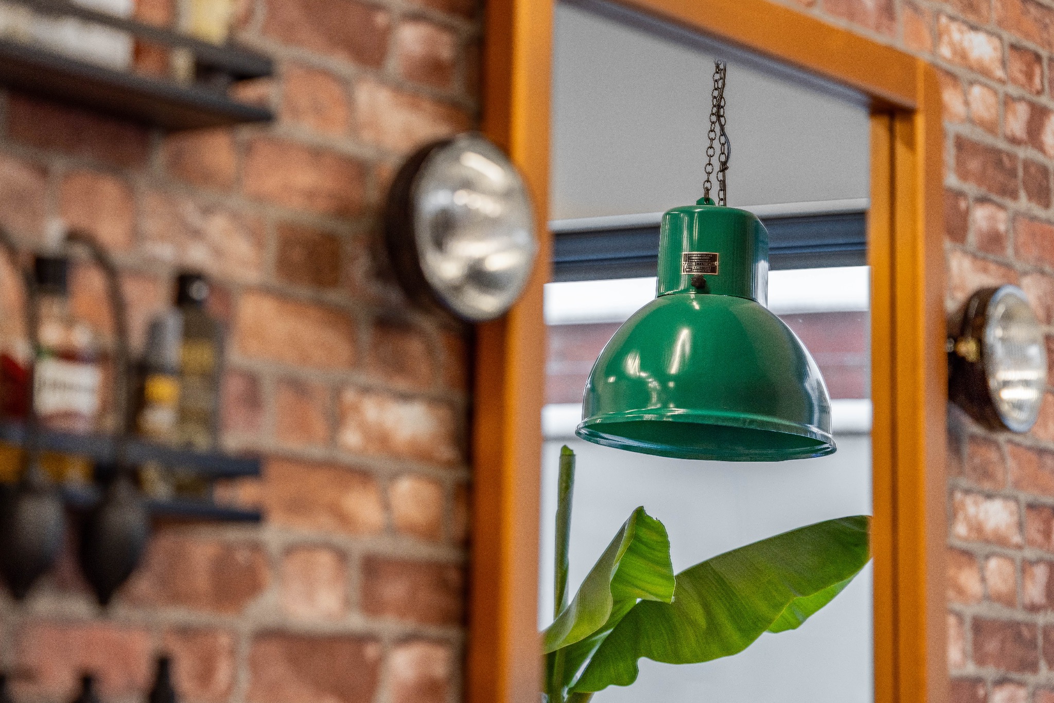 Loftowe wnętrze z ceglaną ścianą, zieloną lampą wiszącą na łańcuchu i reflektorem na ścianie. W tle półki z butelkami. Widok przez ramę lustra.