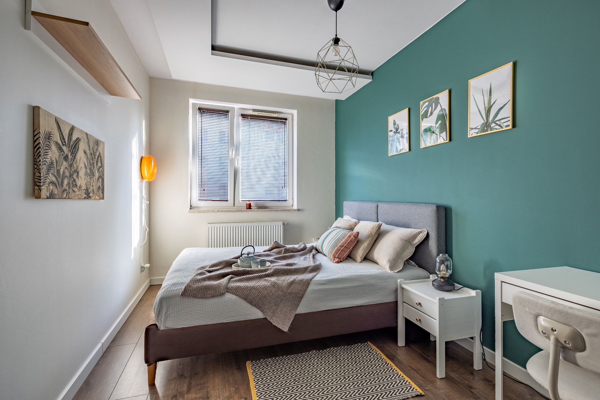 Jasna sypialnia z łóżkiem, stolikiem nocnym i biurkiem. Turkusowa ściana z grafikami roślinnymi. Na łóżku pled, poduszki i zestaw do herbaty. Geometryczna lampa sufitowa.