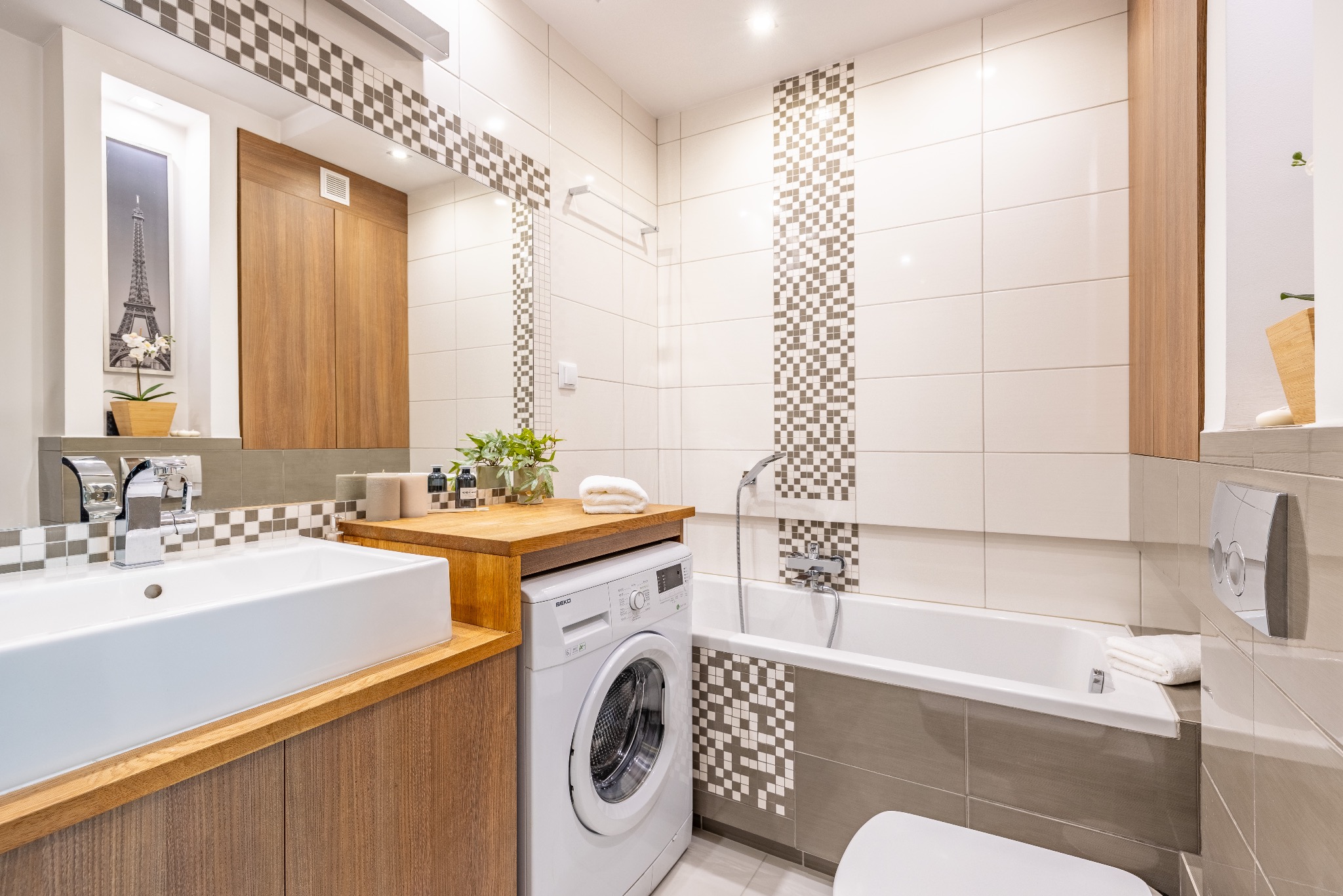 Jasna łazienka z drewnianymi akcentami: umywalka, szafka, blat. Nad umywalką lustro, obok pralka. Na ścianach białe płytki z dekoracyjnym pasem mozaiki.