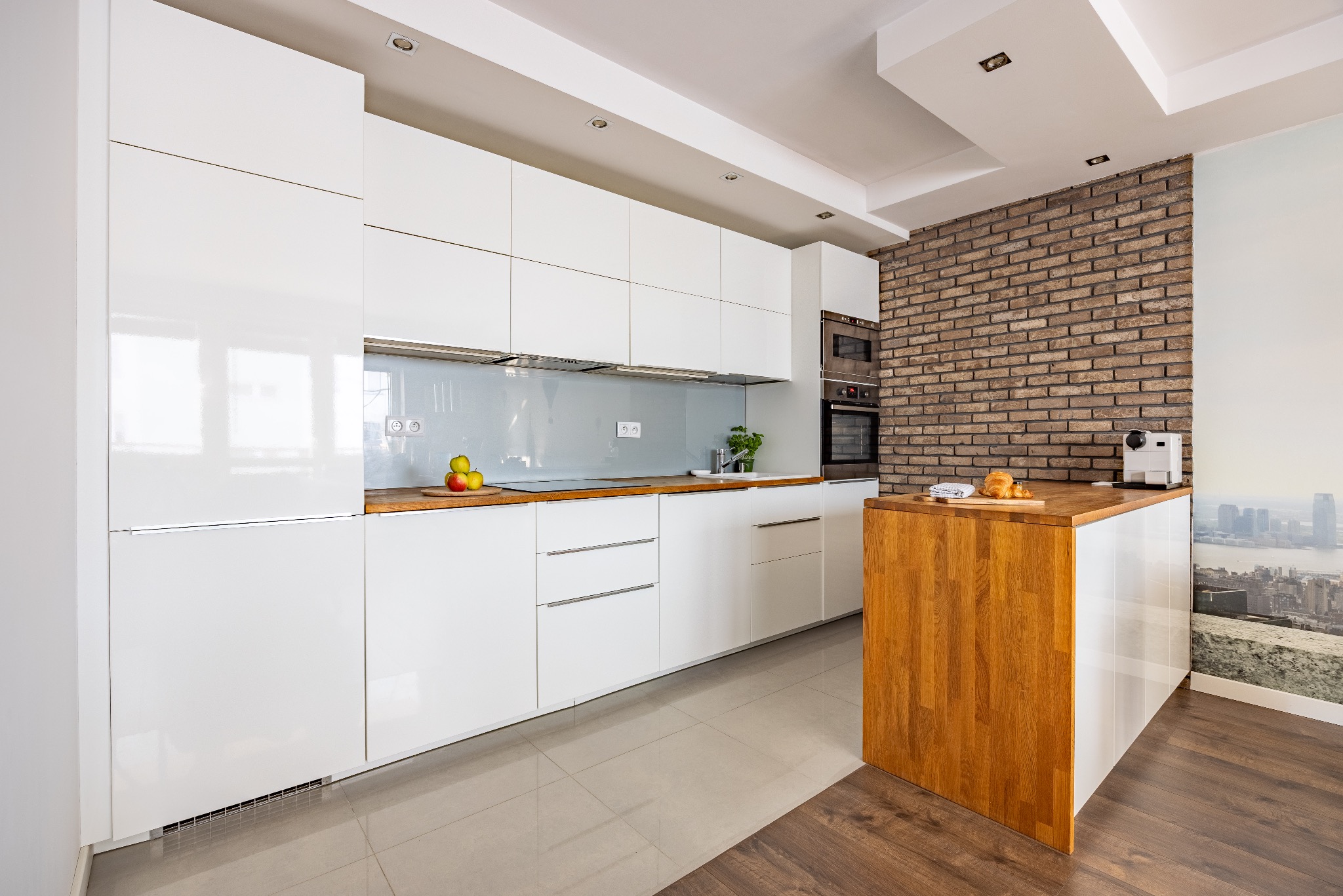 Nowoczesna, jasna kuchnia z białymi, błyszczącymi szafkami i drewnianym blatem. Na blacie owoce, zlew z ziołami. Ceglana ściana i widok na miasto w tle.