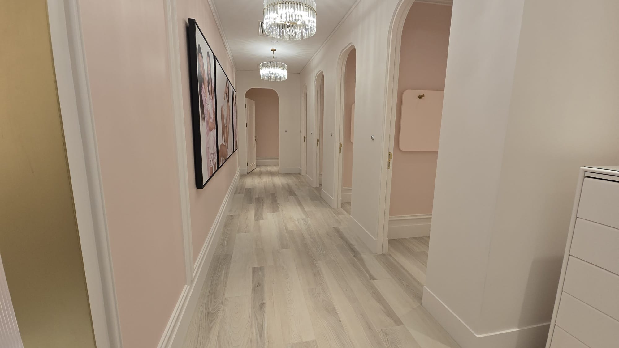 Pastelowy korytarz z drewnianą podłogą, oświetlony kryształowymi lampami. Na ścianie wiszą oprawione grafiki, a w tle widoczne są łukowe przejścia.