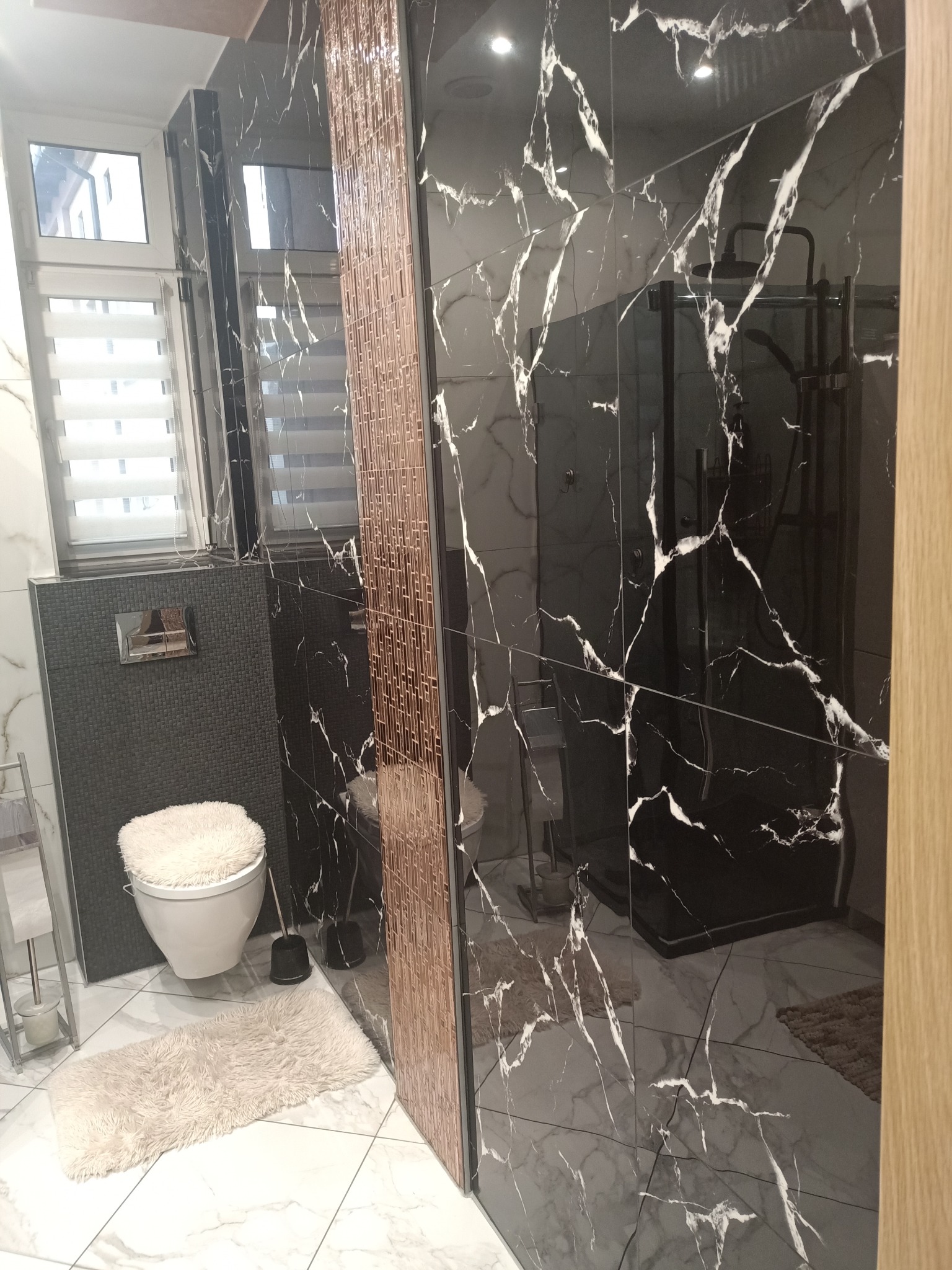 Nowoczesna łazienka z czarnymi płytkami imitującymi marmur, kabiną prysznicową typu walk-in i toaletą z puszystą nakładką. Widoczne detale wykończeniowe i oświetlenie.