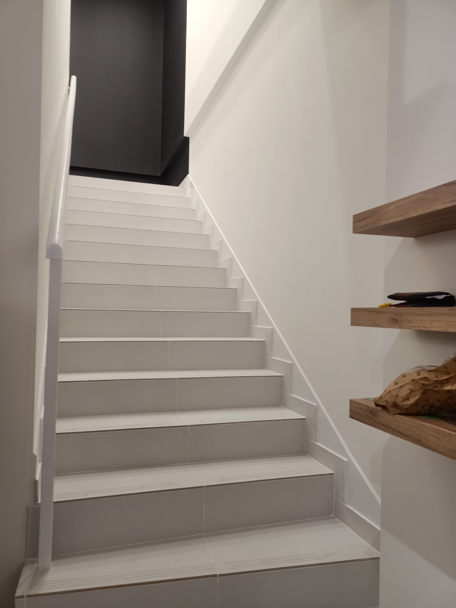 Białe, wykafelkowane schody z minimalistyczną balustradą i drewnianymi półkami na ścianie. Nowoczesne wykończenie wnętrza z akcentem na detale.