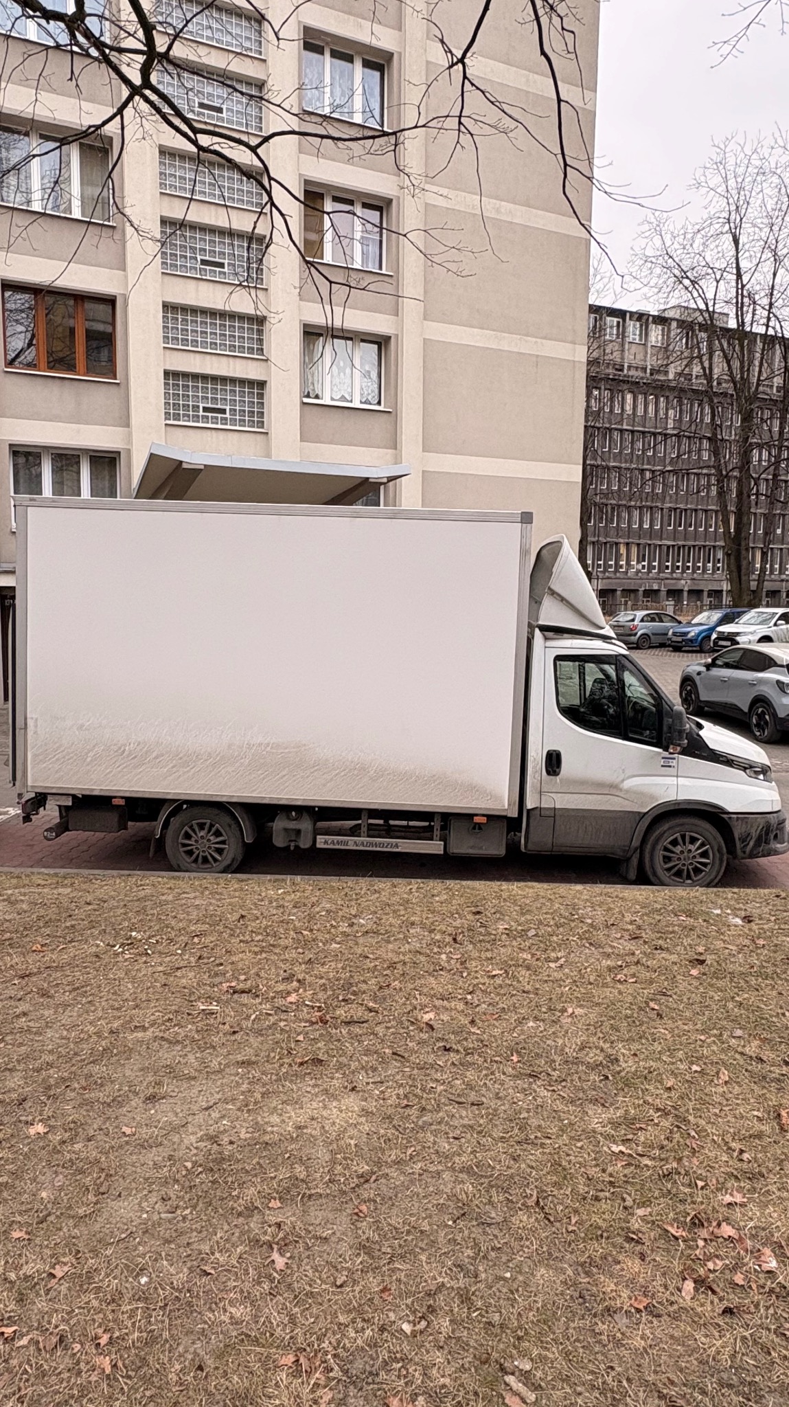 Biały samochód dostawczy z zabudową kontenerową, zaparkowany na trawniku przed blokiem mieszkalnym w pochmurny dzień. Widok z boku, na drugim planie budynki i zaparkowane auta.