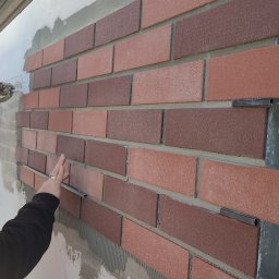 Norbert Grzegorczyk - Montaż dekoracyjnej okładziny ściennej z cegiełek w odcieniach czerwieni i brązu. Widoczne metalowe elementy mocujące i dłoń osoby układającej. Ściana w trakcie prac.