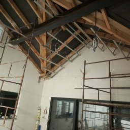 Norbert Grzegorczyk - Skośny sufit z drewnianą konstrukcją i metalowym stelażem pod płyty. Widoczne rusztowania i fragment osoby. Prace wykończeniowe w jasnym pomieszczeniu.