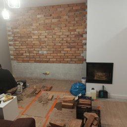 Norbert Grzegorczyk - Wnętrze w trakcie prac: mur z cegły klinkierowej na ścianie, widoczny kominek, narzędzia i materiały budowlane, osoba w trakcie pracy. Ujęcie z perspektywy podłogi.