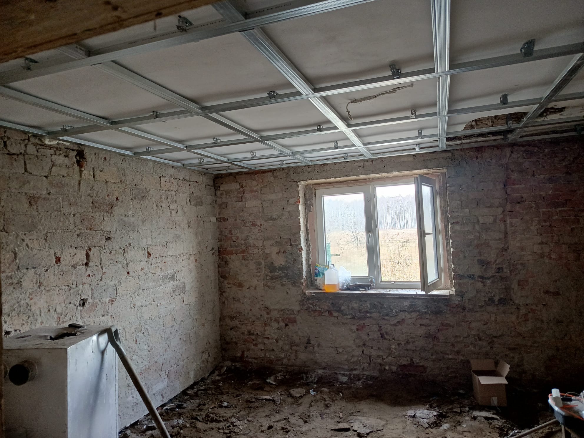 Surowy pokój z odsłoniętą cegłą i metalową konstrukcją sufitu podwieszanego. Widoczne narzędzia i resztki materiałów budowlanych. Okno z widokiem na zewnątrz.