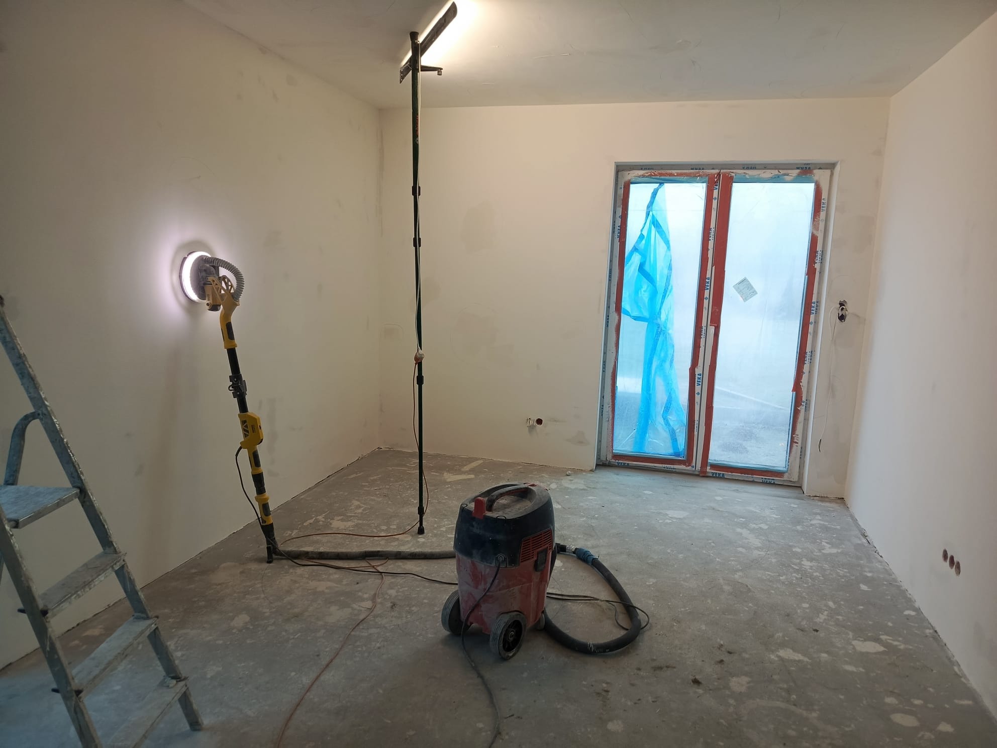 Wnętrze pokoju w trakcie szpachlowania: szlifierka do gładzi, odkurzacz przemysłowy, drabina i okno zabezpieczone folią. Widoczne ślady szpachli na ścianach i podłodze.