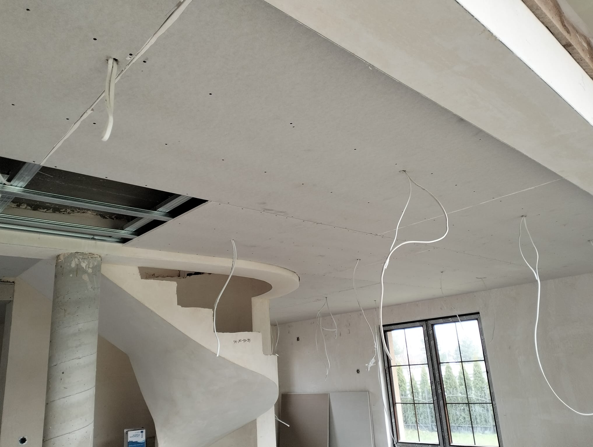 Sufit z płyt gipsowo-kartonowych w trakcie montażu, widoczne kable elektryczne i fragment spiralnych schodów. Surowy stan wykończenia wnętrza.