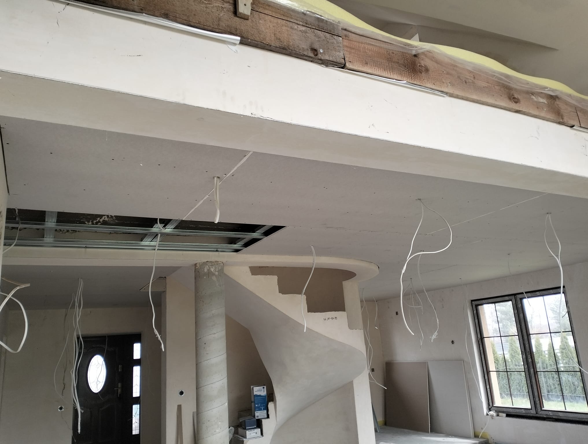 Wnętrze w trakcie prac wykończeniowych: sufit z płyt karton-gips, widoczne kable, kolumna ze schodami spiralnymi. Surowy stan, przygotowanie do dalszych prac.