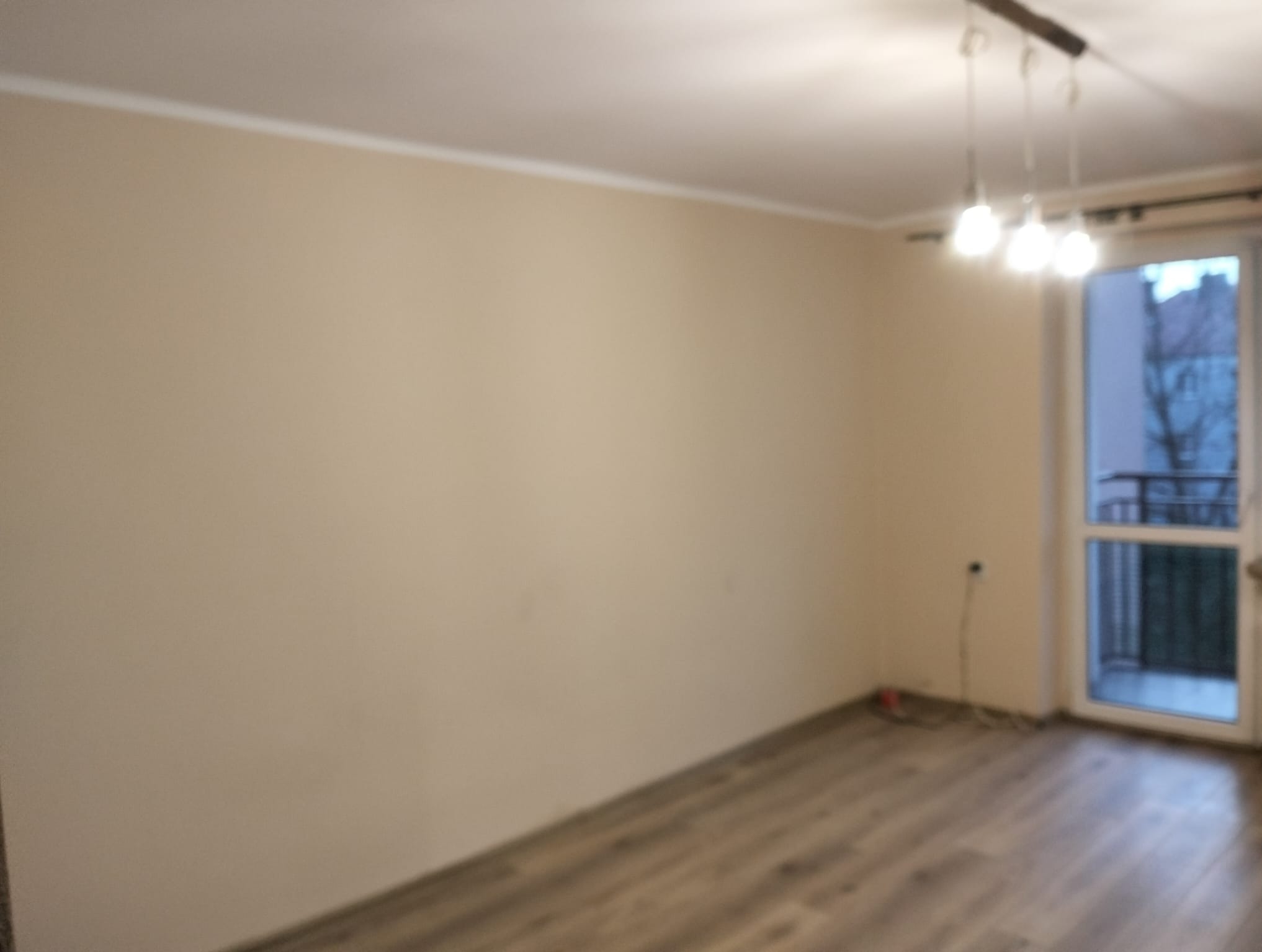 Jasne wnętrze pokoju z panelami podłogowymi i świeżo pomalowanymi ścianami w odcieniach beżu. Widoczne listwy przypodłogowe i sufitowe oraz lampa sufitowa.