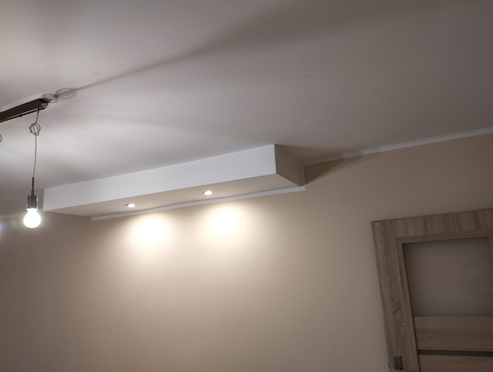 Biała zabudowa sufitowa z oświetleniem punktowym w beżowym pokoju, widoczna lampa wisząca i fragment drzwi. Minimalistyczny design, wykończenie wnętrz.