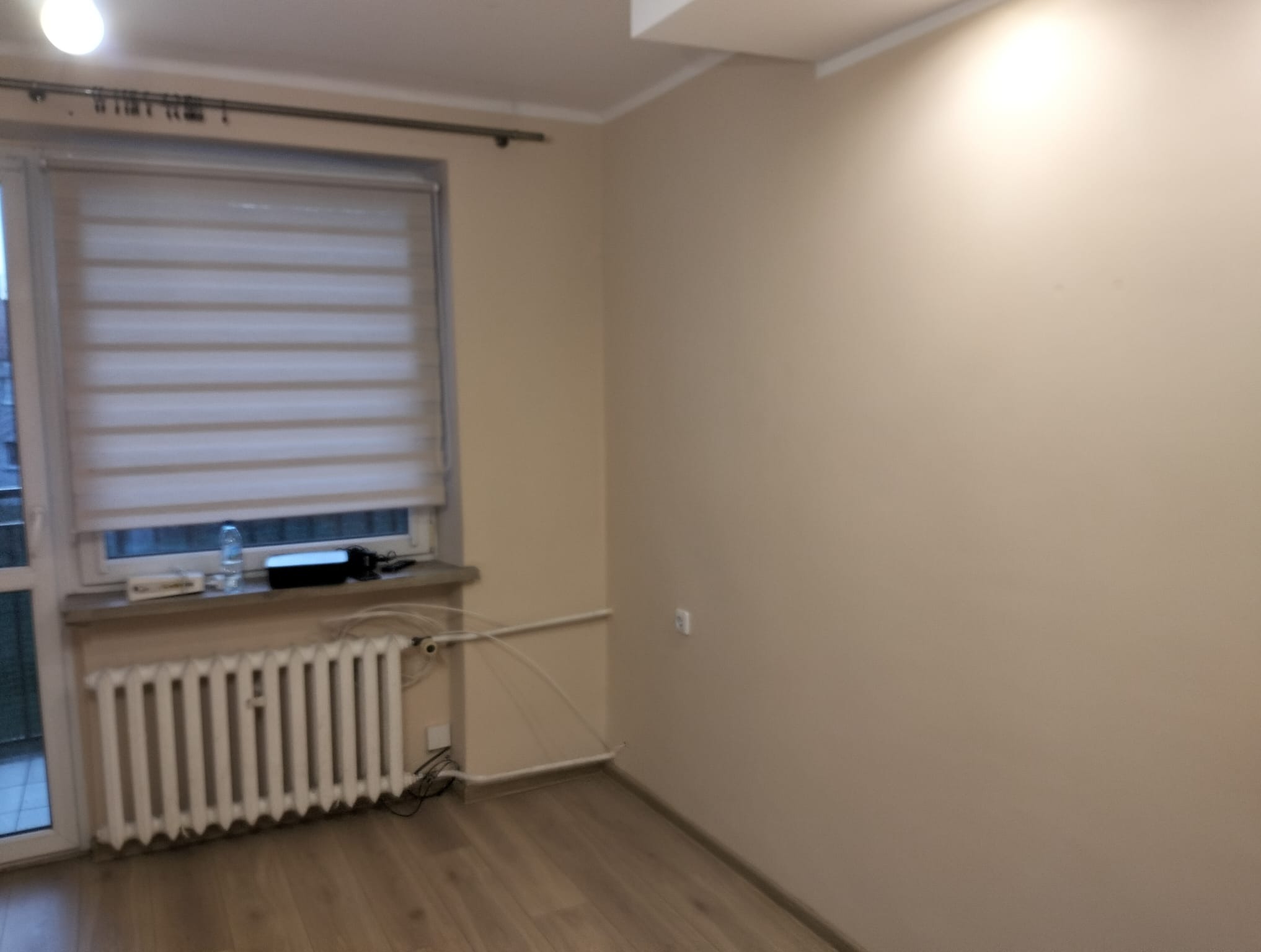 Jasne wnętrze pokoju po odświeżeniu: beżowe ściany, drewnopodobna podłoga, biały grzejnik pod oknem z roletami. Widoczny fragment sufitu i gniazdko elektryczne.