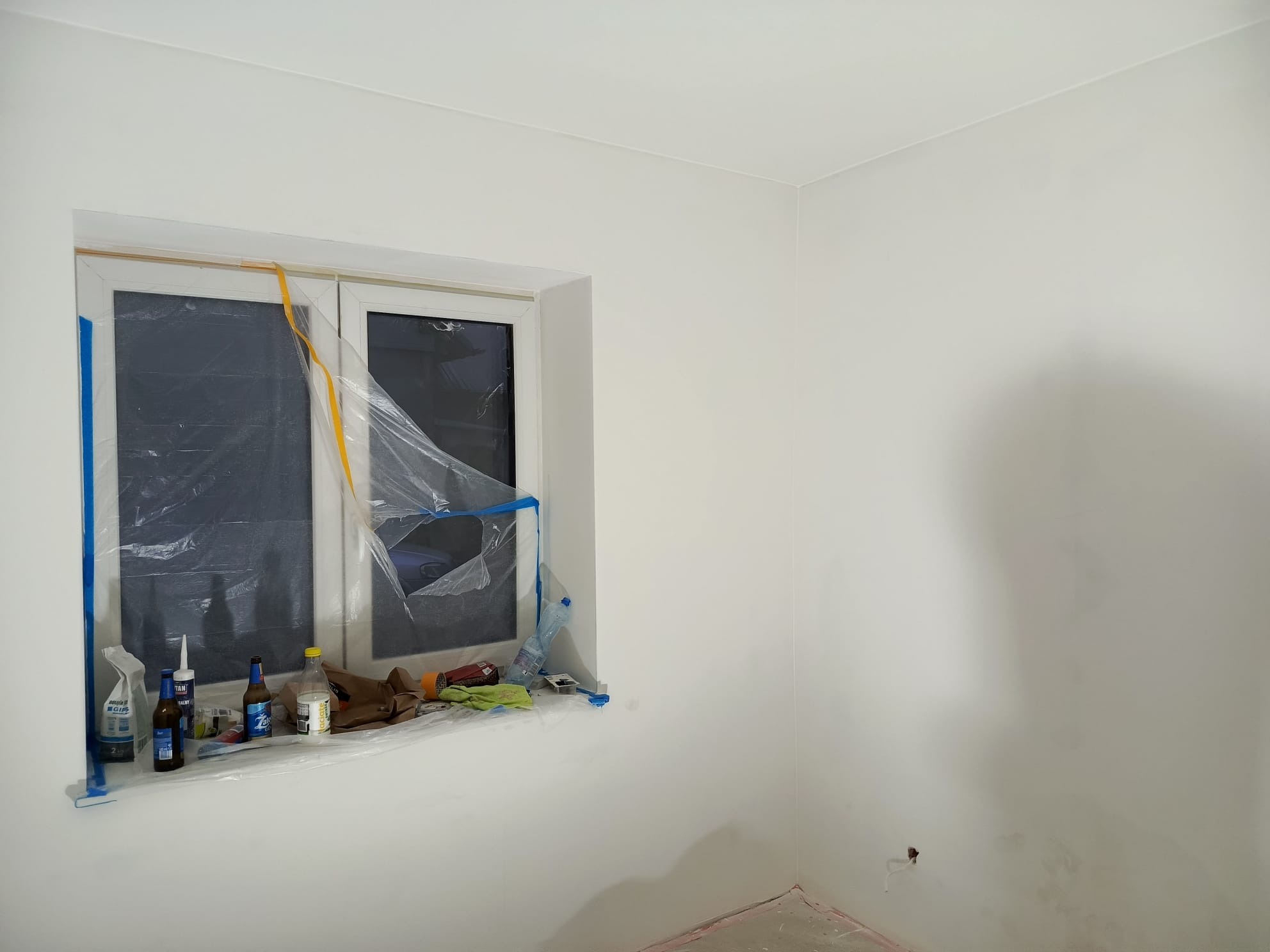 Białe, świeżo pomalowane wnętrze pokoju z oknem zabezpieczonym folią malarską. Na parapecie widoczne akcesoria malarskie i butelki. Ściany gładkie, bez dekoracji.