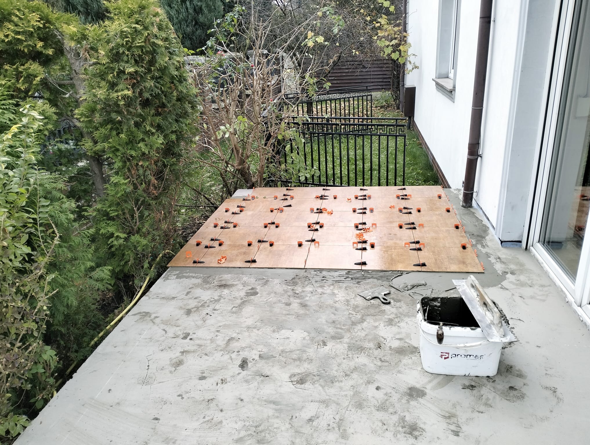 Układanie płytek na tarasie z użyciem systemu poziomowania. Widoczny pojemnik z zaprawą i narzędzia. Tło: zieleń i biała elewacja budynku.