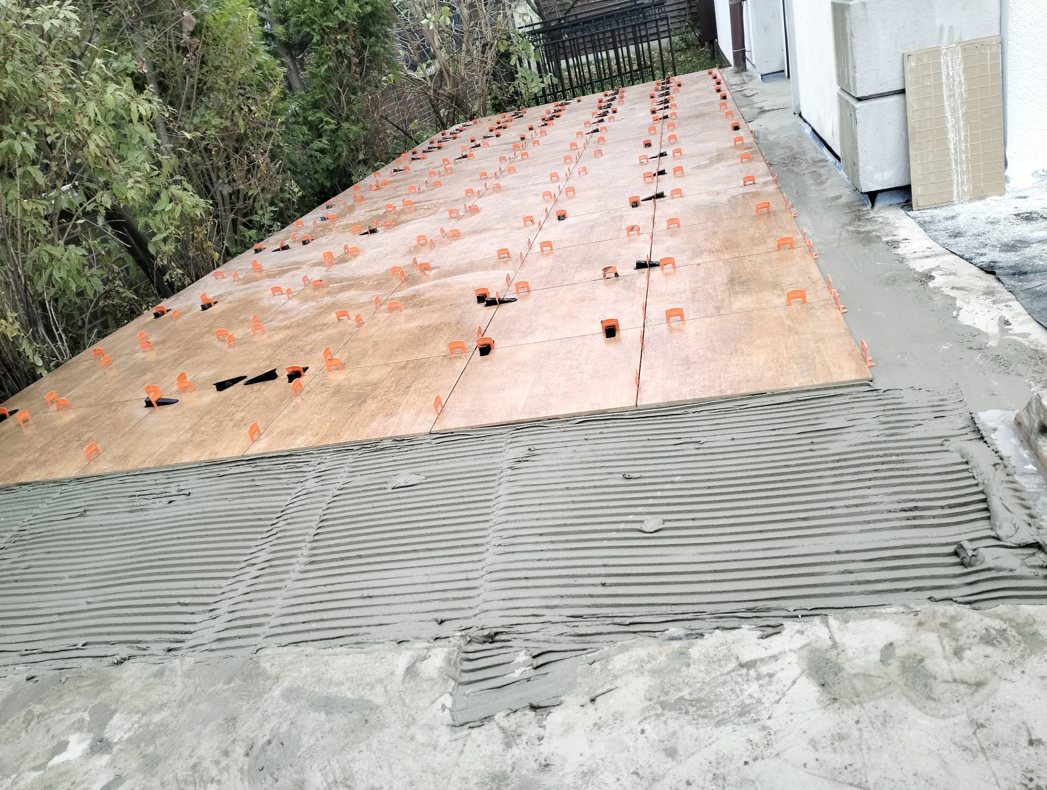 Układanie płytek na zewnątrz z użyciem klipsów poziomujących. Widoczna warstwa kleju z charakterystycznymi rowkami i częściowo ułożone płytki w ciepłych odcieniach brązu.