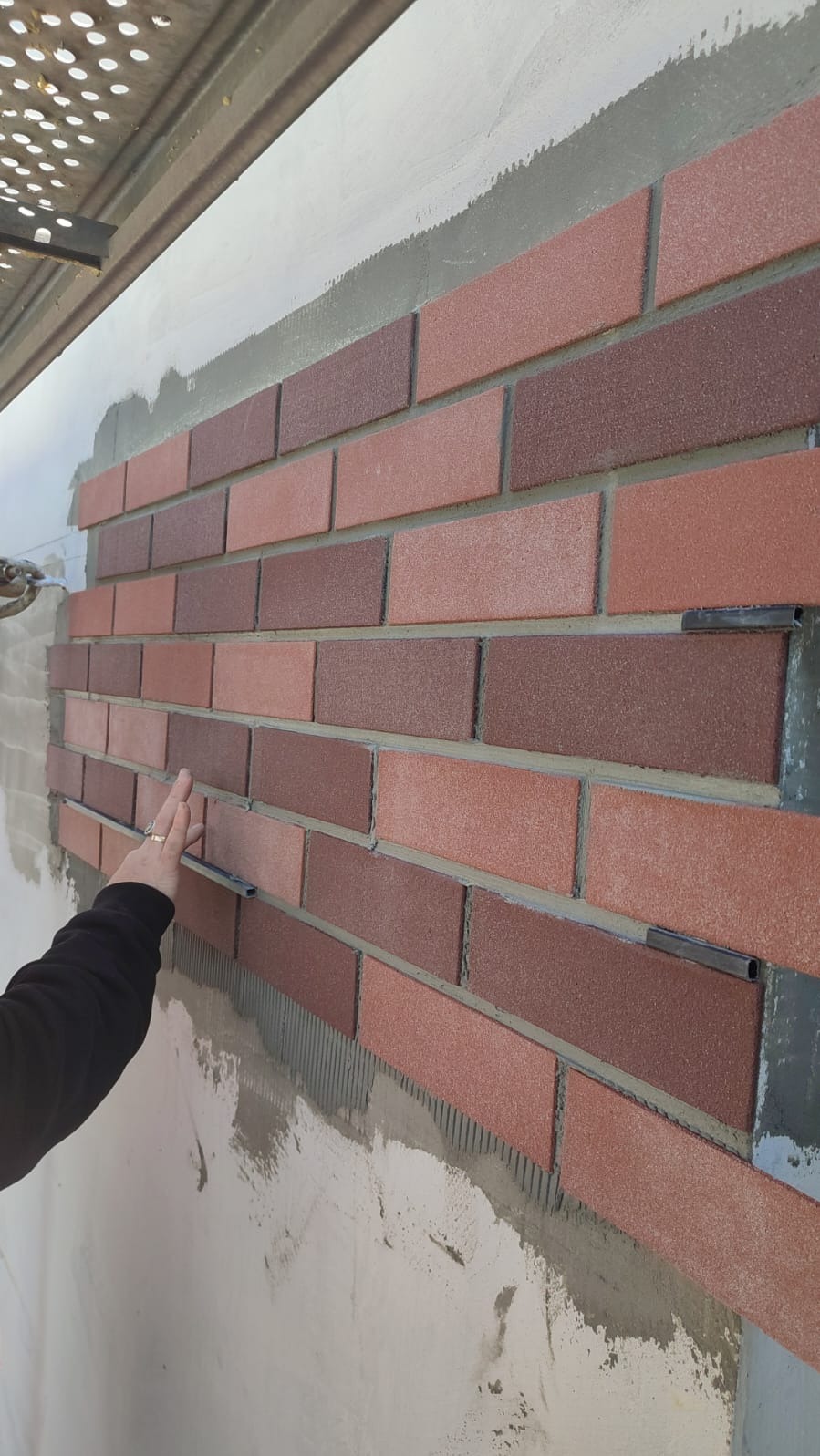 Montaż dekoracyjnej okładziny ściennej z cegiełek w odcieniach czerwieni i brązu. Widoczne metalowe elementy mocujące i dłoń osoby układającej. Ściana w trakcie prac.