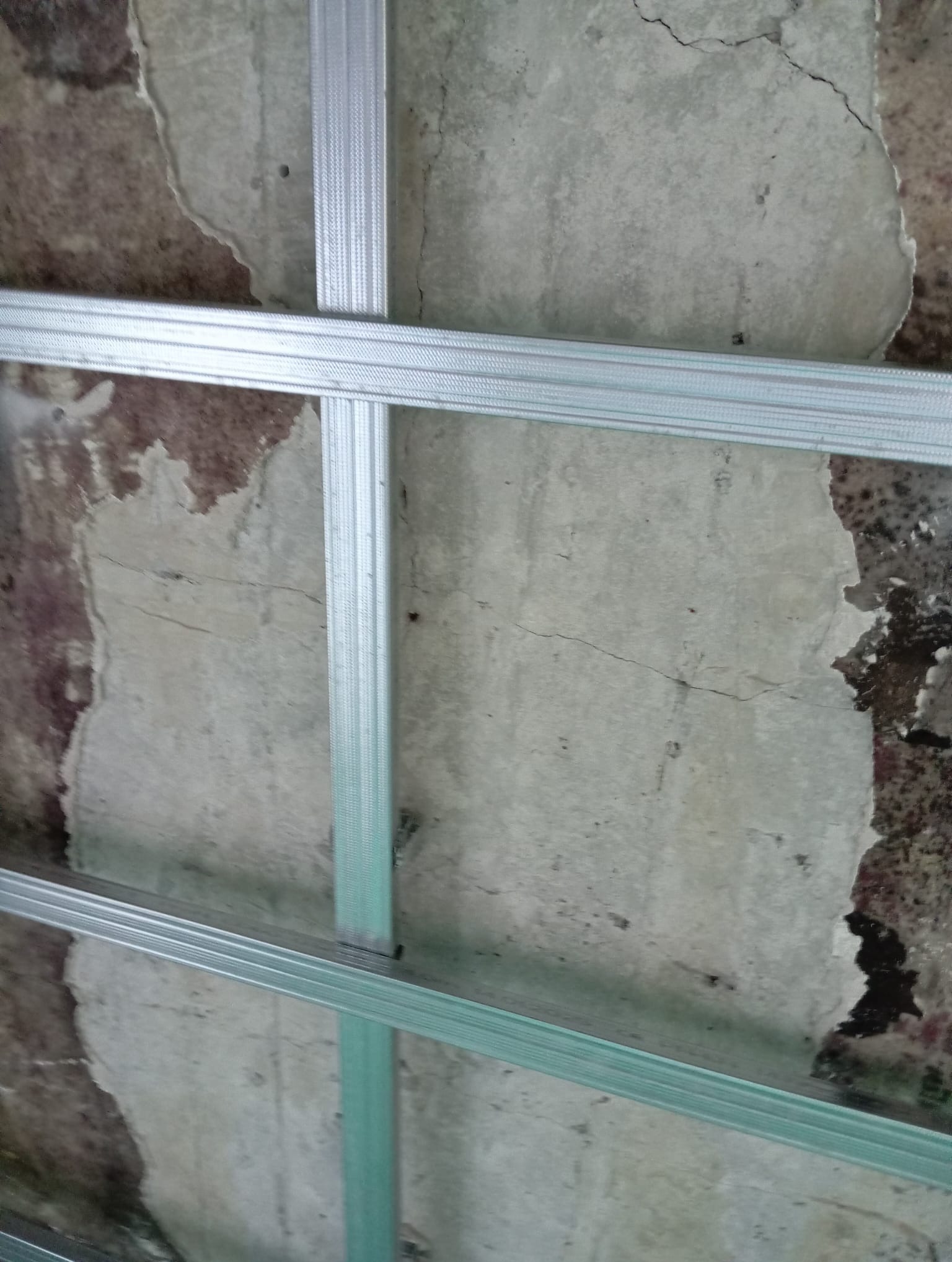 Metalowy stelaż sufitowy na tle starego, popękanego tynku. Widoczne ślady po odpadającym malowaniu i nierówności powierzchni. Ujęcie z dołu, podkreślające kontrast faktur.