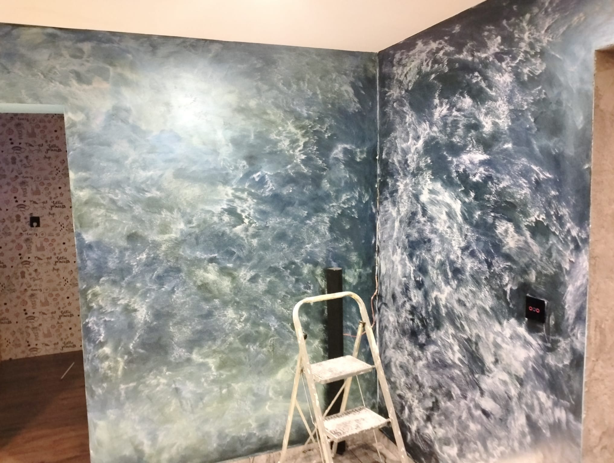 Dekoracyjna ściana w odcieniach błękitu i bieli, imitująca wzburzone morze. Obok drabina malarska i fragment ściany z tapetą w dziecięcym pokoju. Efektowna technika malarska.