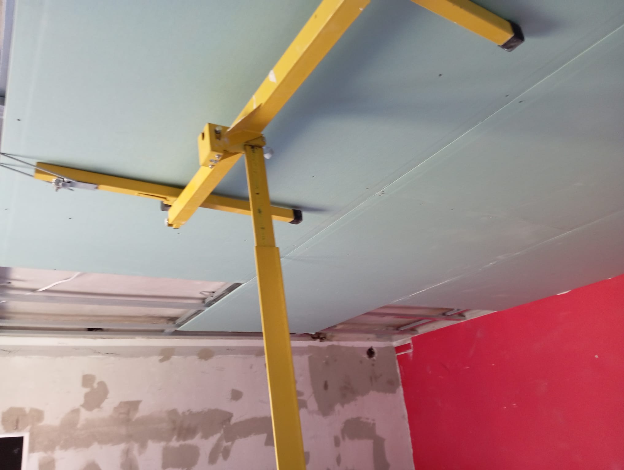 Żółty podnośnik do płyt G-K podtrzymuje szarą płytę na suficie. Widoczne fragmenty ściany w stanie surowym i pomalowanej na czerwono.